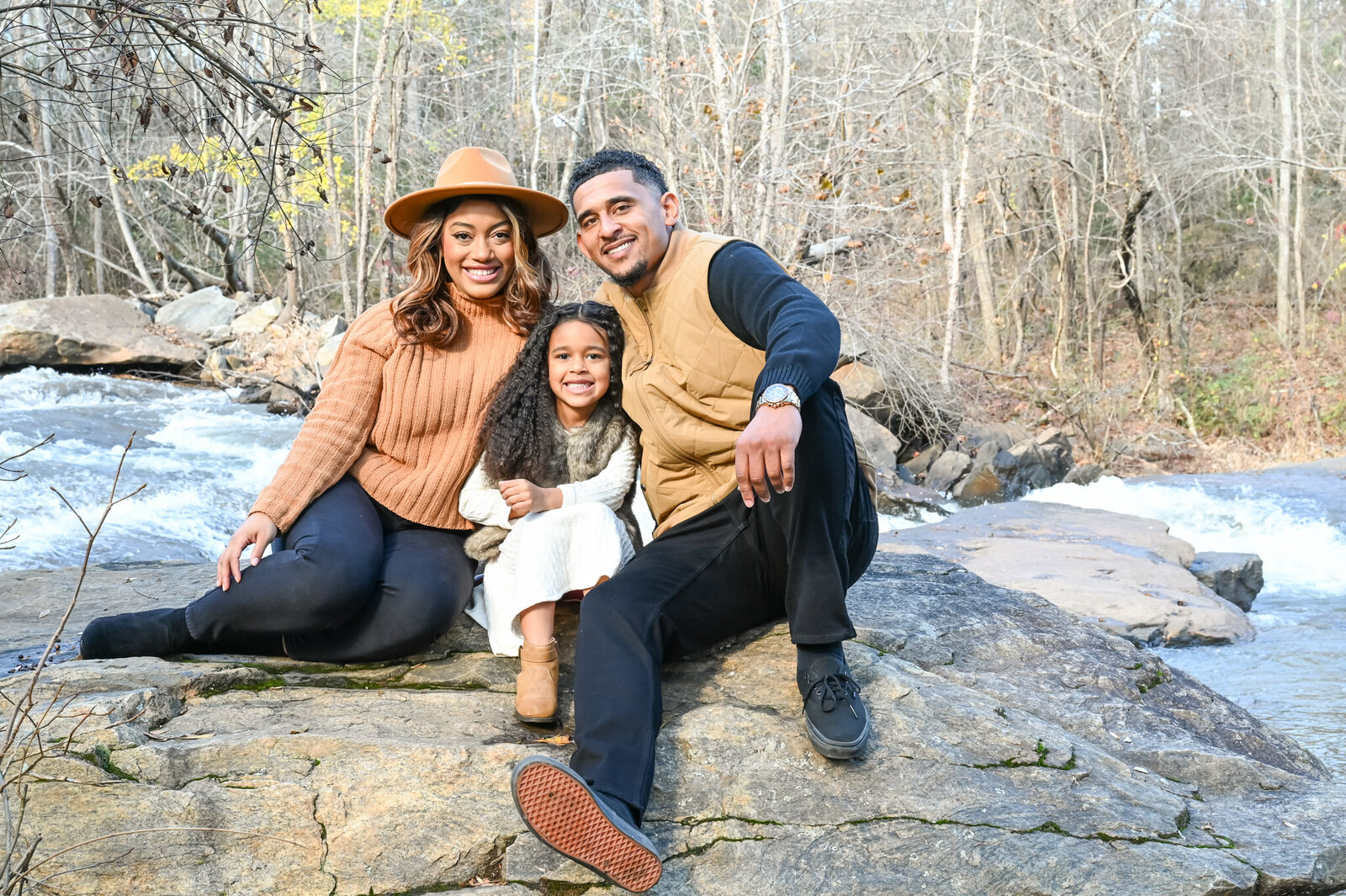 a family of mother father and daughter sitting on rocks smiling at the camera photographed by Millz Photography in Greenville, SC