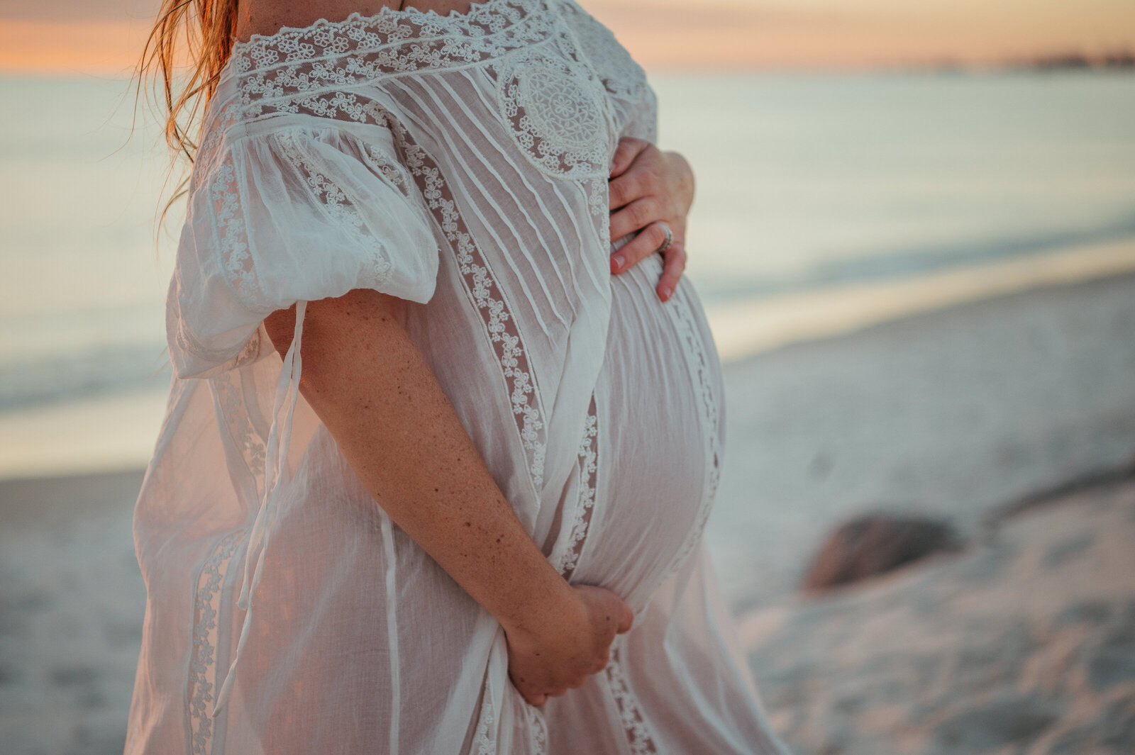 Close up photo of pregnant belly in a flowing white dress at sunset on the beach