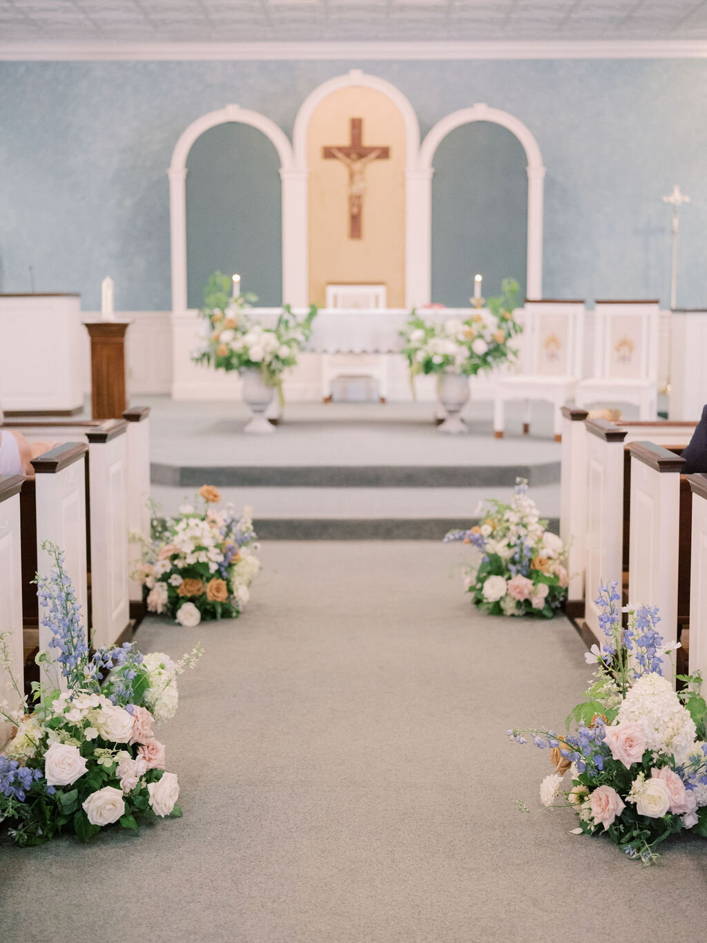 Ceremony in Sacred Heart Catholic Church in Chestertown MD with ground arrangements going down the aisle and two large arrangements in urns at the altar with florals such as blue delphinium, white hydrangea, blush roses, white dahlias and toffee roses.