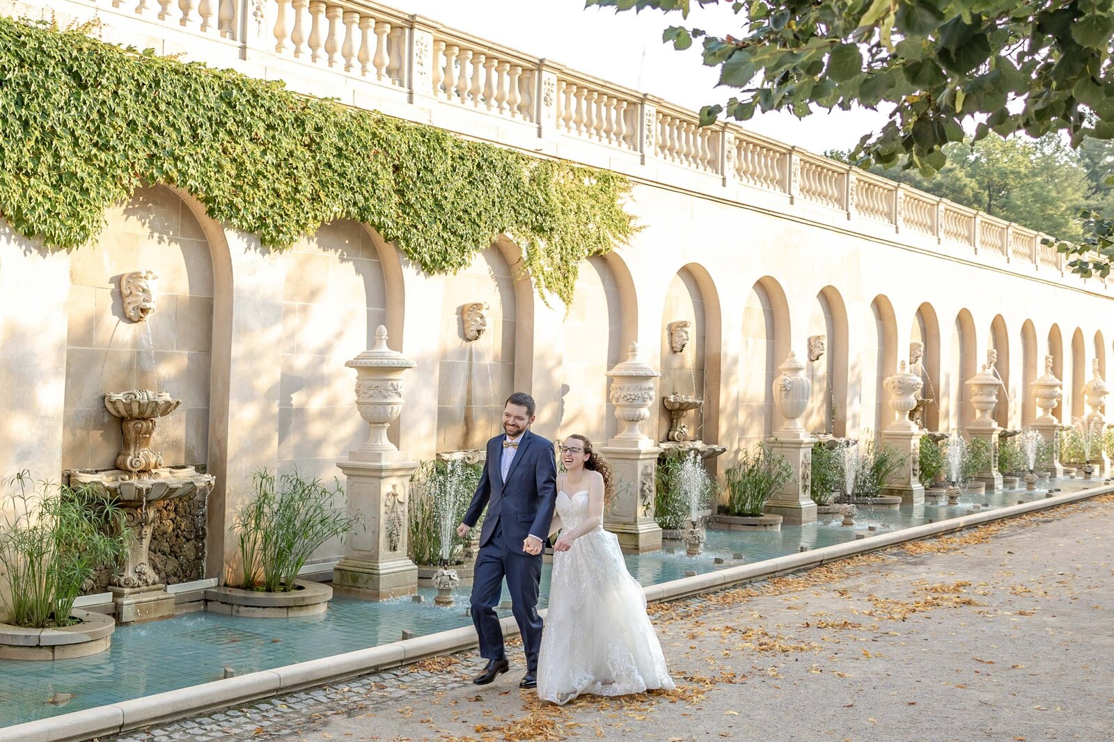 Bride and groom walking hand in hand along a picturesque fountain at their wedding venue Longwood Gardens, capturing the romance of their special day in Kennett Square, Pennsylvania.