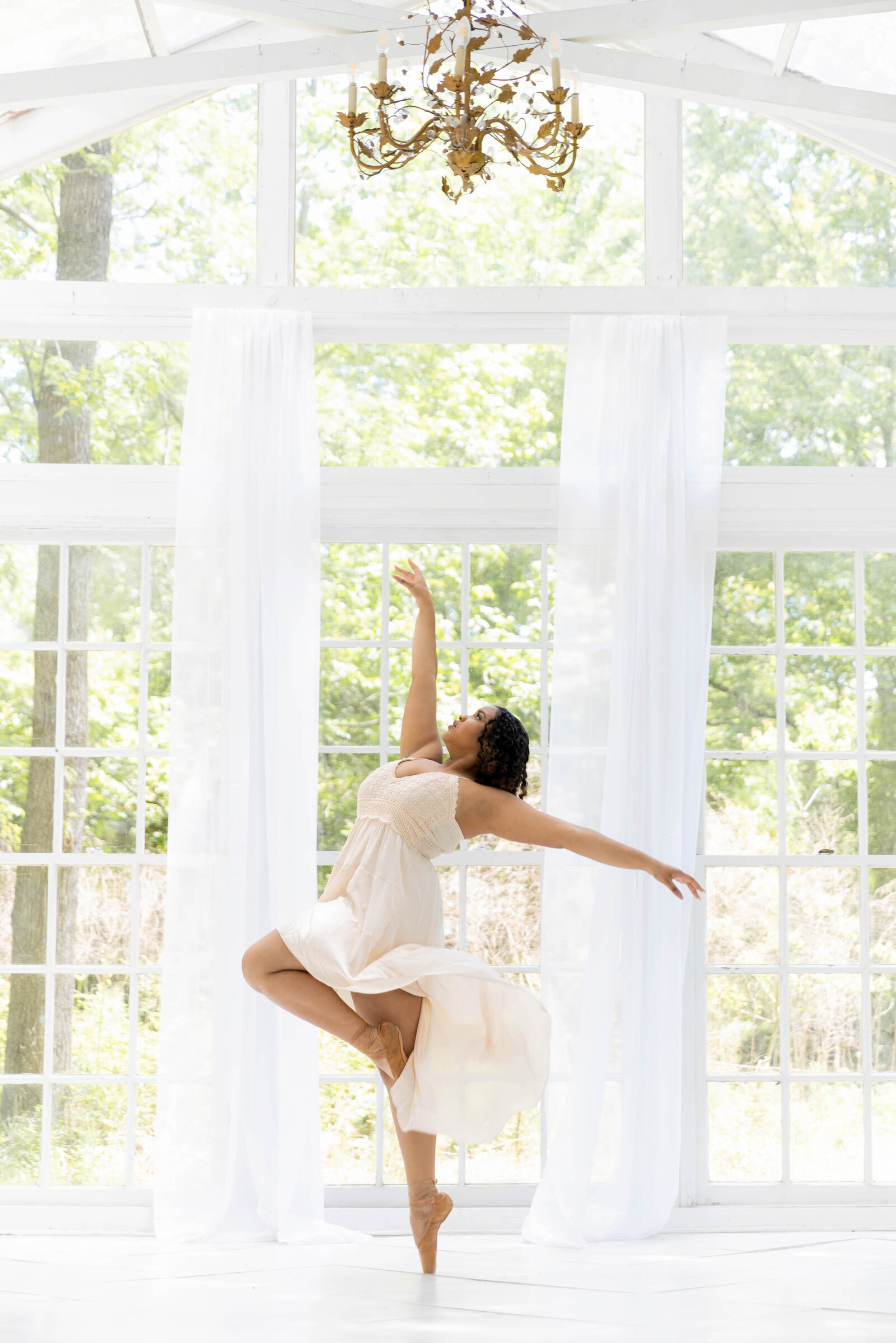 Black ballerina in white dress and brown pointe shoes dancing in glass greenhouse under a brass chandelier