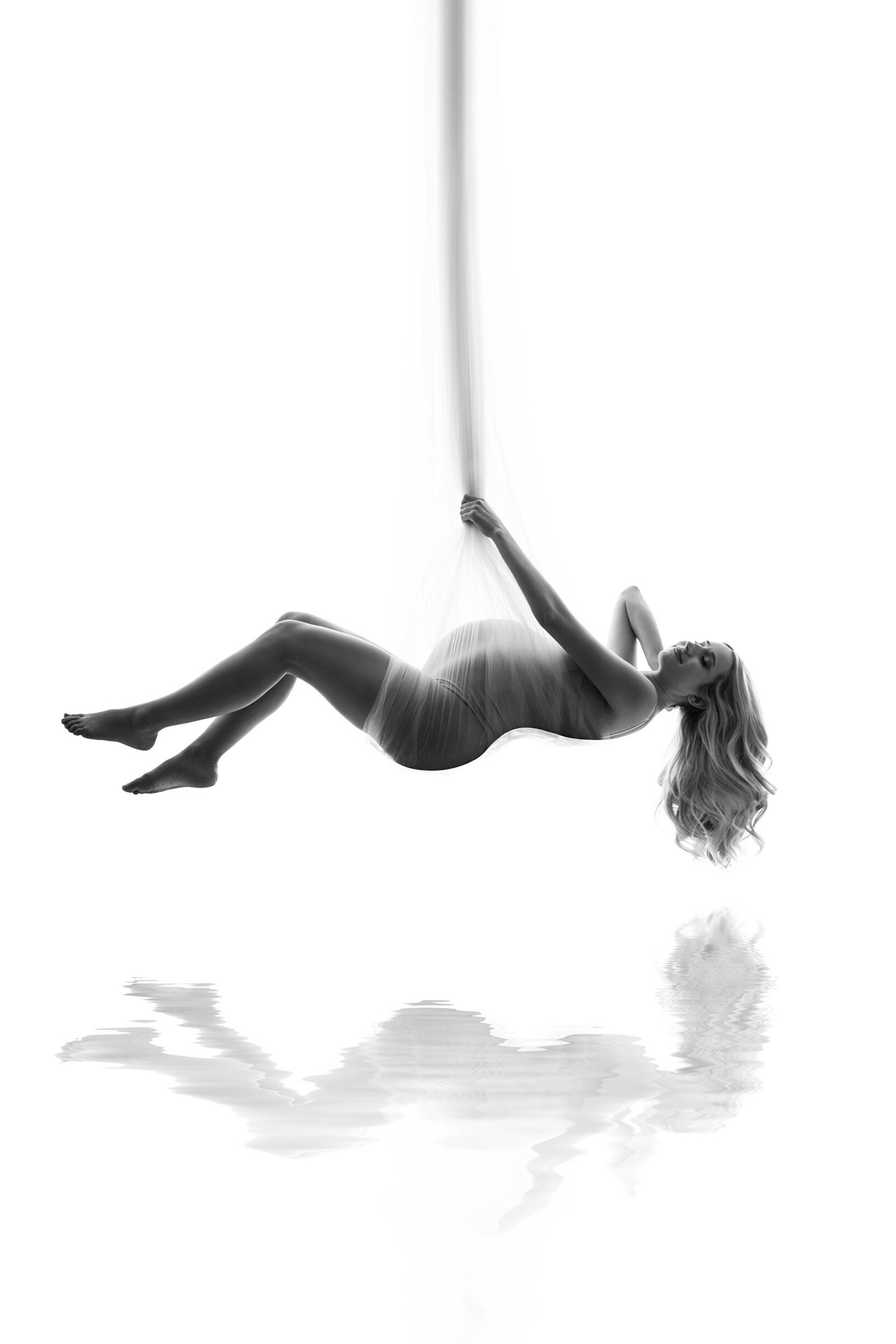 A black and white photo of a pregnant woman in our Waukesha studio suspended in sheer fabric over water.