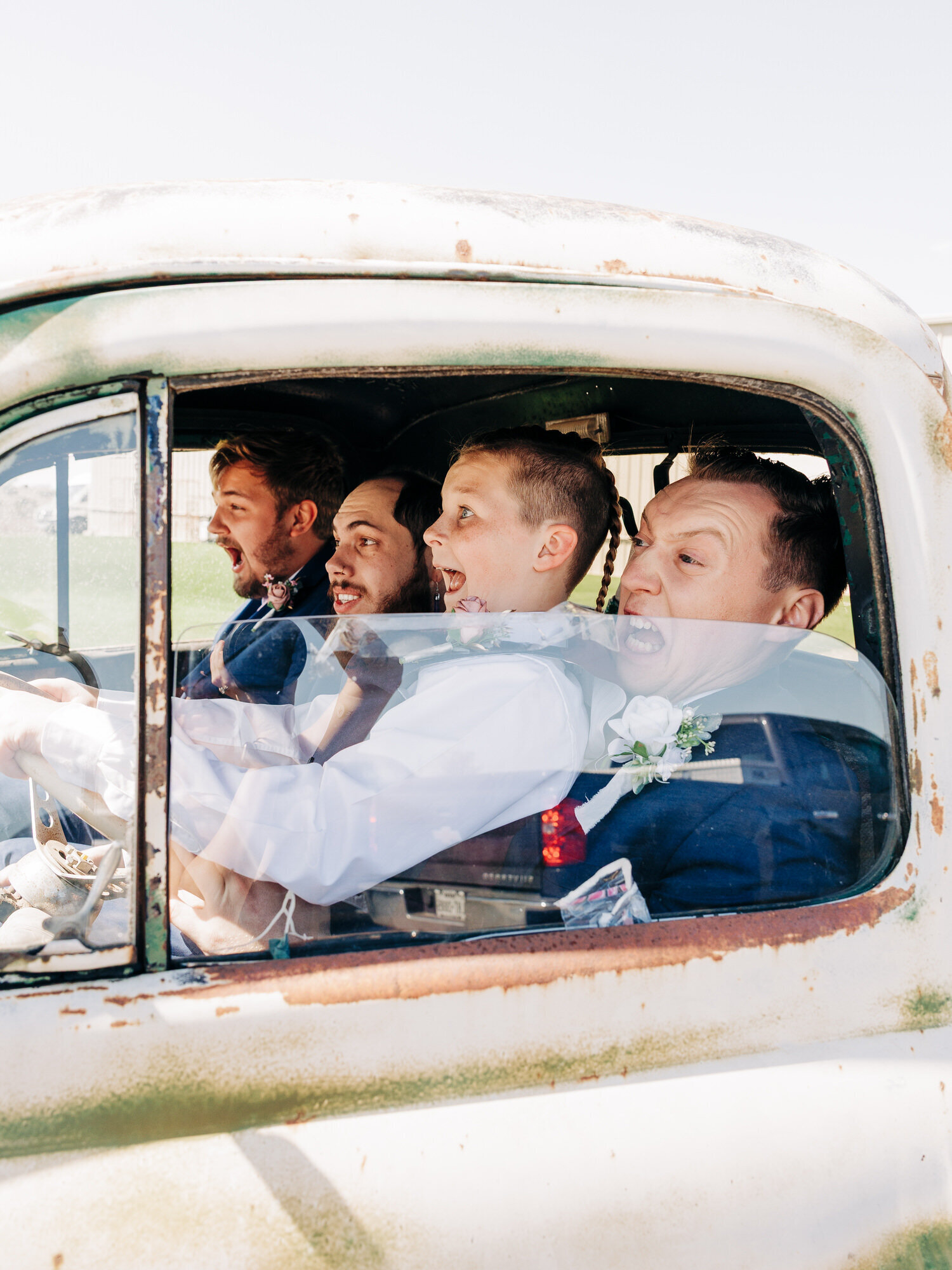 Four men dressed in formal attire sit in an old, rustic truck, smiling and laughing. Captured by an Eau Claire wedding photographer, the drivers excitement and sunlight reflecting their joy make for a memorable, heartwarming moment.



