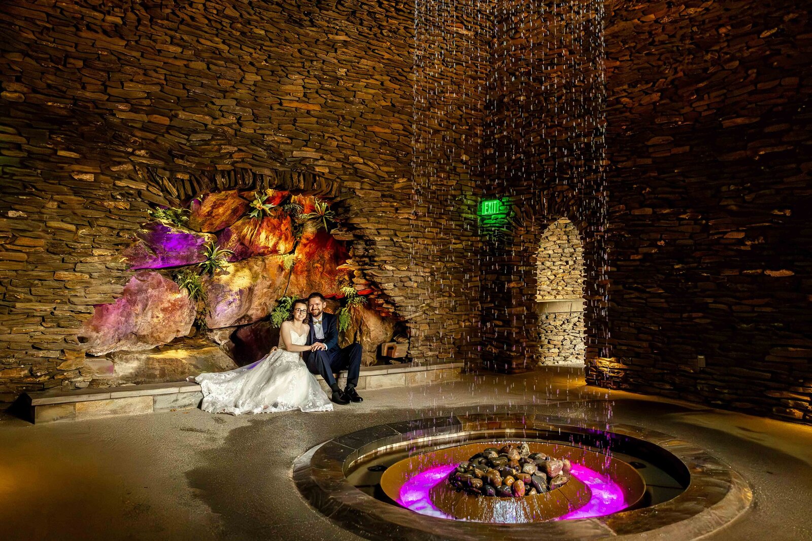 The bride and groom share a romantic moment in a stone-walled room with a cascading waterfall and glowing purple lights, captured during a Longwood Gardens wedding in beautiful Kennett Square,PA blending rustic elegance with unique charm.