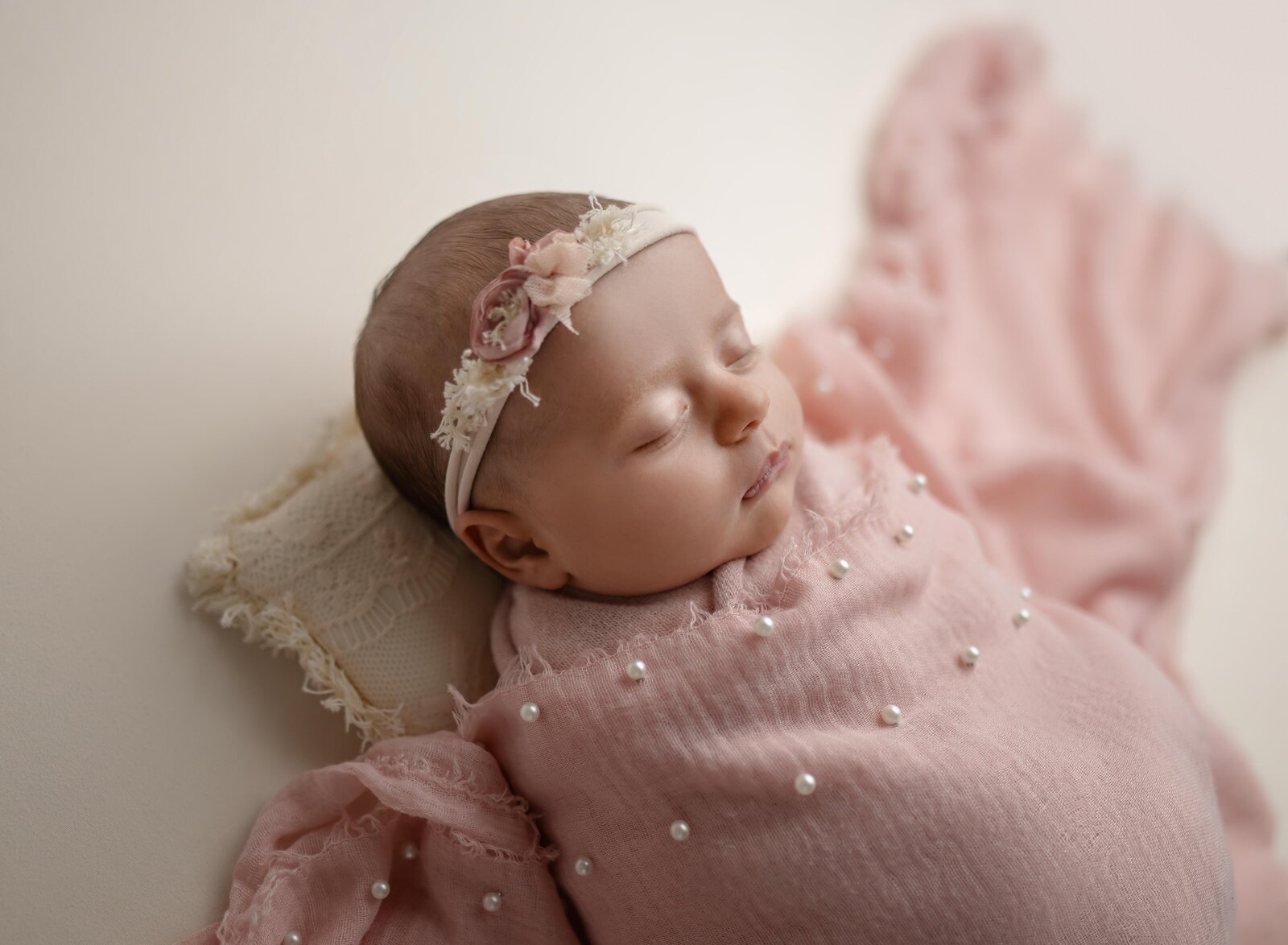 Baby girl sleeping on a cream pillow with a pink pearl wrap over her.