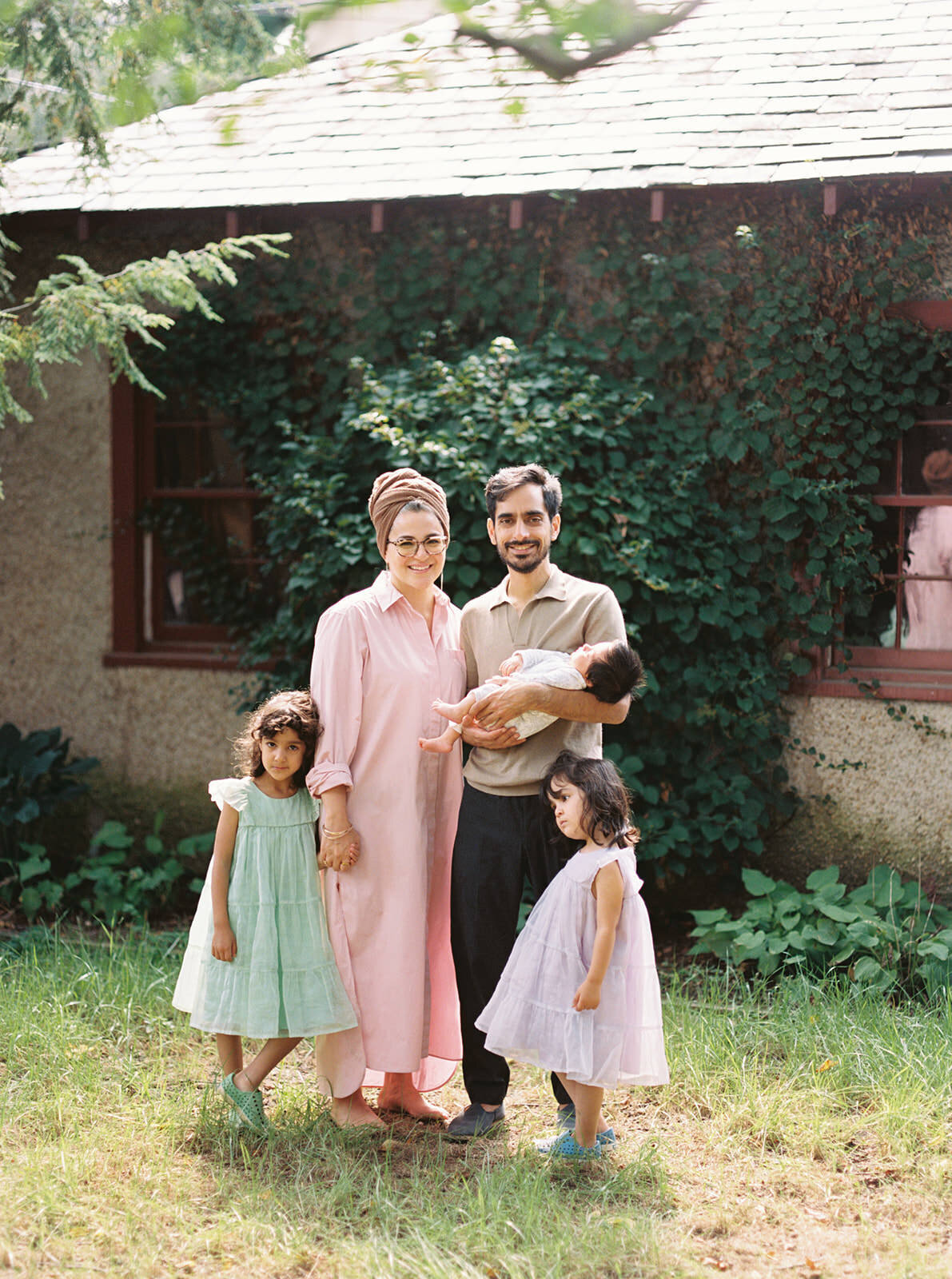 Family of five posing outdoors in front of a vine-covered house, showcasing gentle smiles.