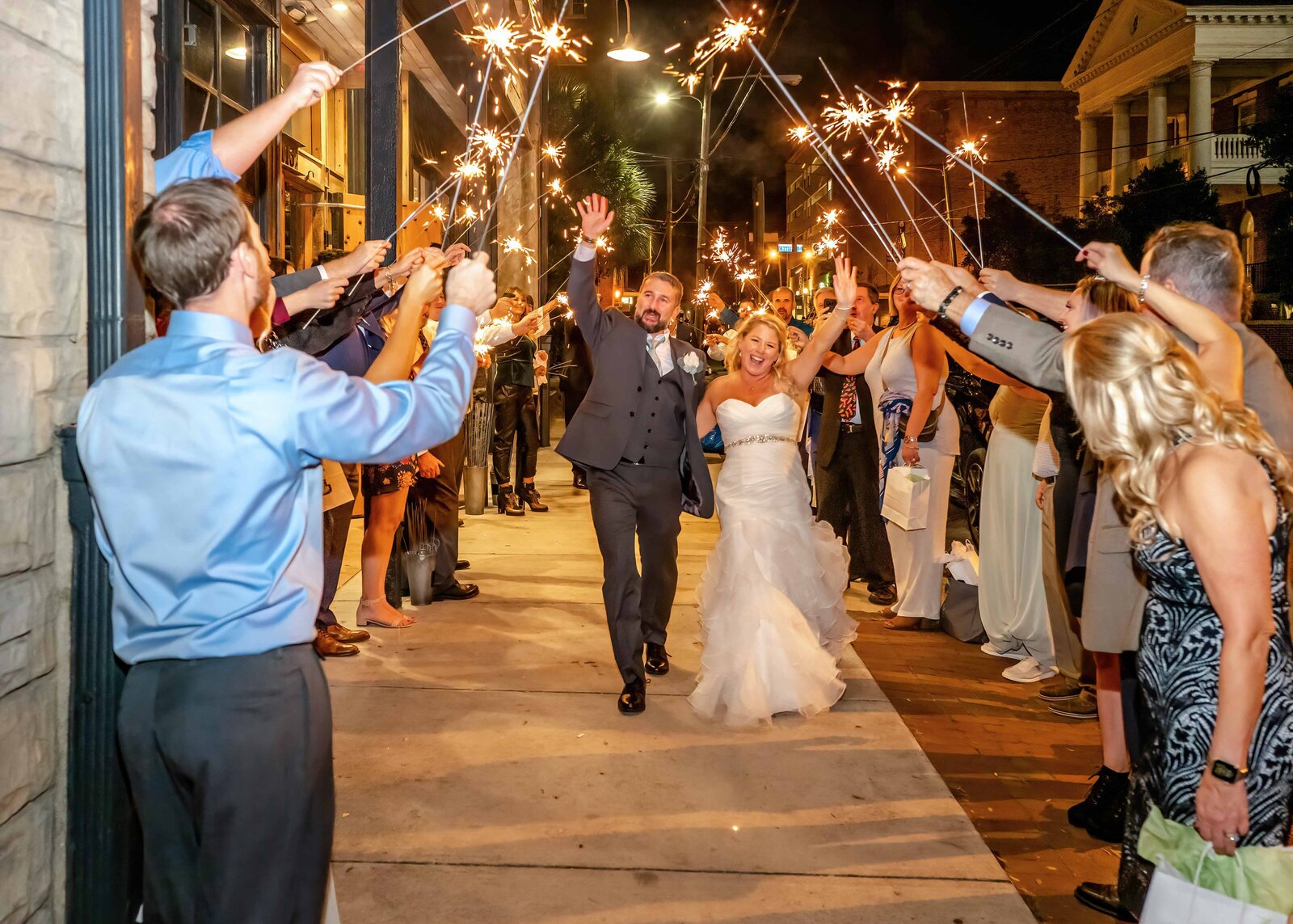 The bride and groom celebrate with a sparkler exit on a downtown Wilmington, NC street, surrounded by cheering guests, blending urban charm with wedding day excitement.