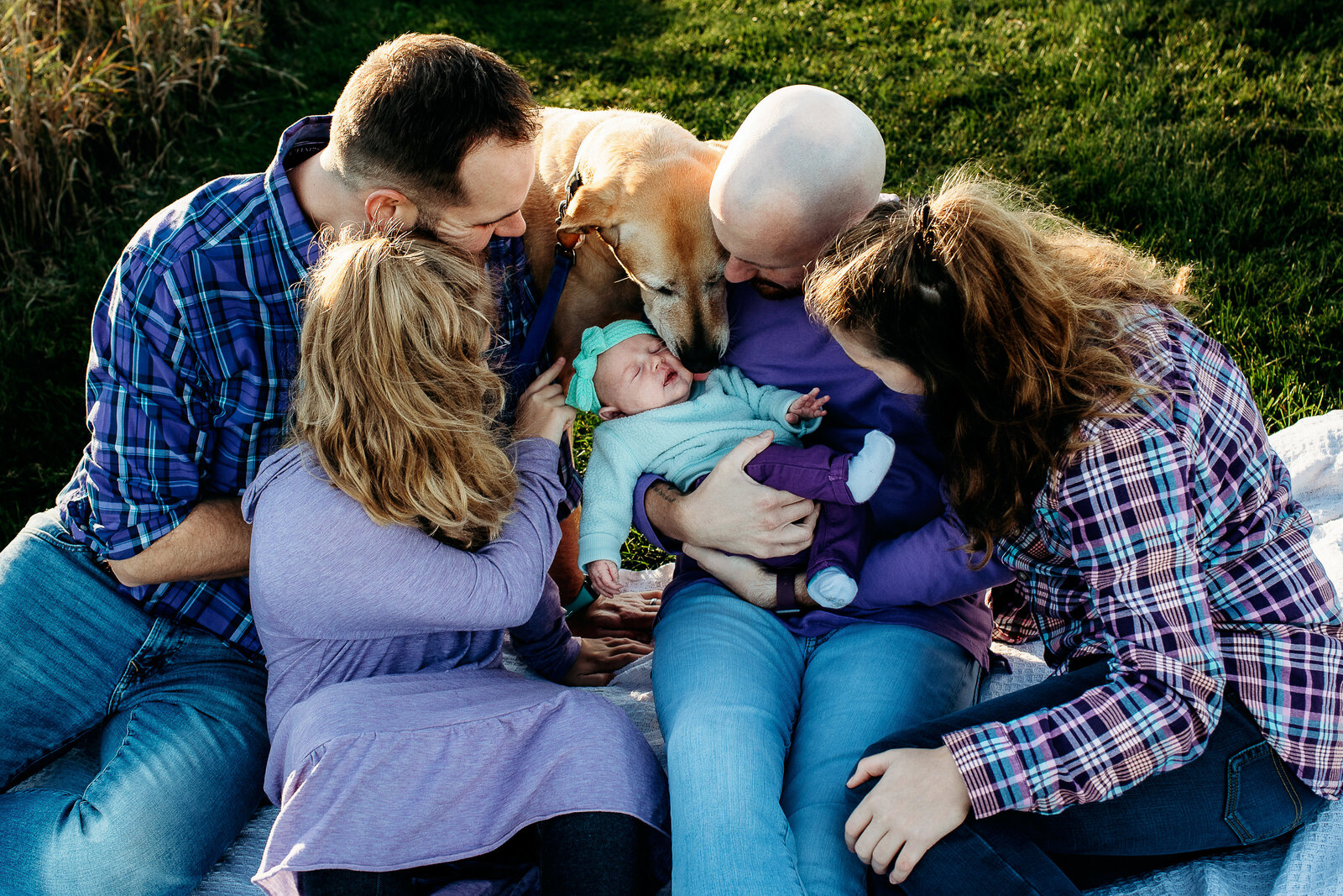 Dads and their two older children snuggling their new baby, their dog peeking over to lick her too.