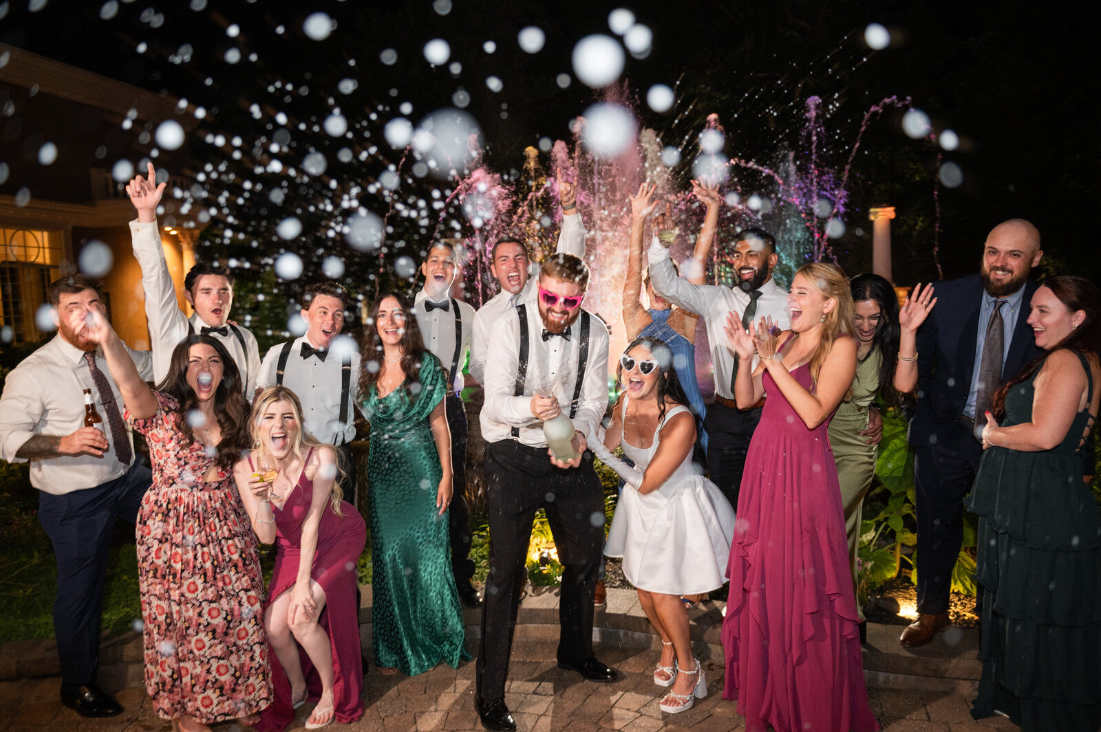 Group of guests and a bride an groom spraying champagne in front of a colorful fountain at night at Brigalias in South Jersey.