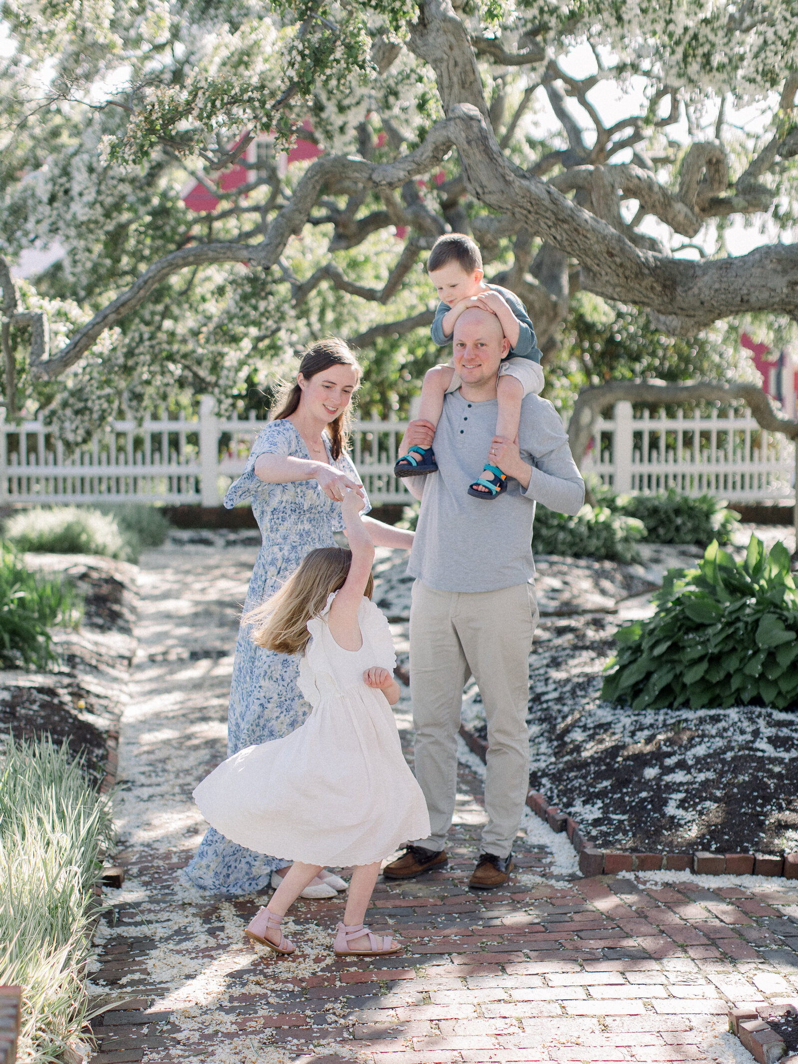 Family of four in a spring garden under flowering cherry trees, mom spinning daughter while dad holds son on his shoulders by NH newborn photographer Fieldstone Studio.