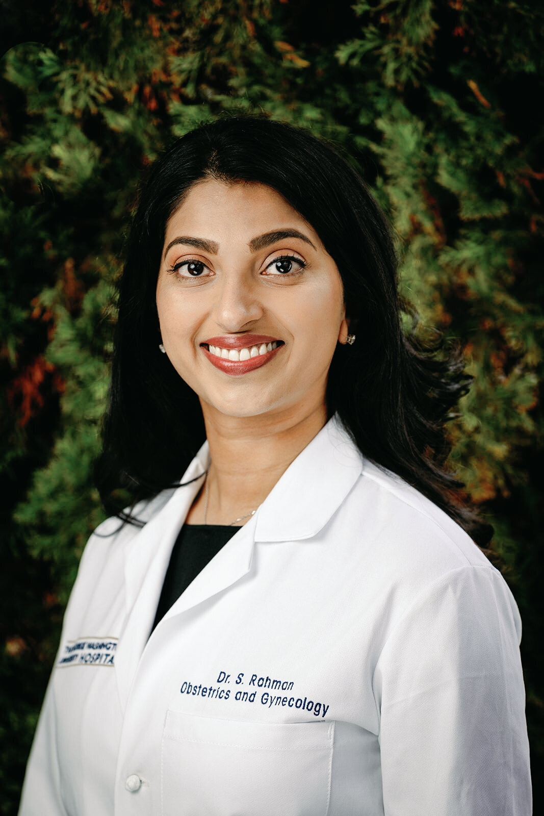 Professional headshot of a female doctor wearing a white coat with her name embroidered, smiling in front of a natural, green backdrop.