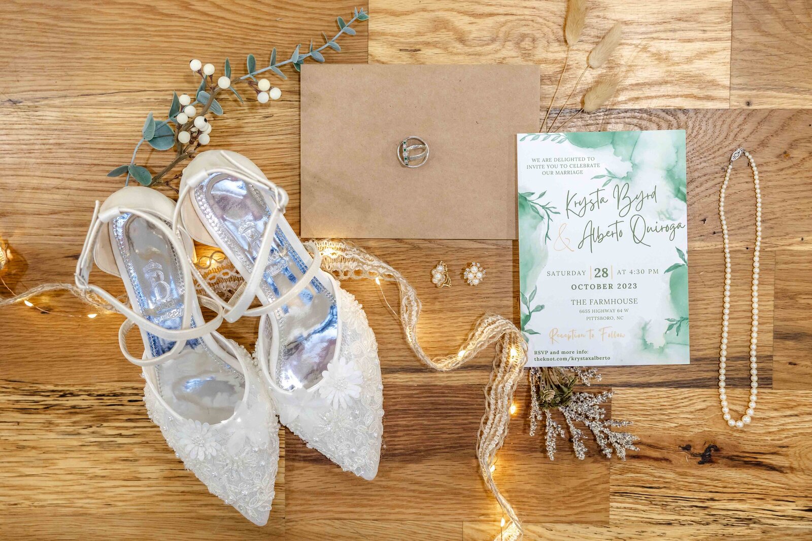 A bridal flat lay featuring lace wedding shoes, a greenery-themed invitation, pearl jewelry, and soft florals, captured on wooden flooring during a Wilmington, NC wedding, blending elegant and natural wedding details.