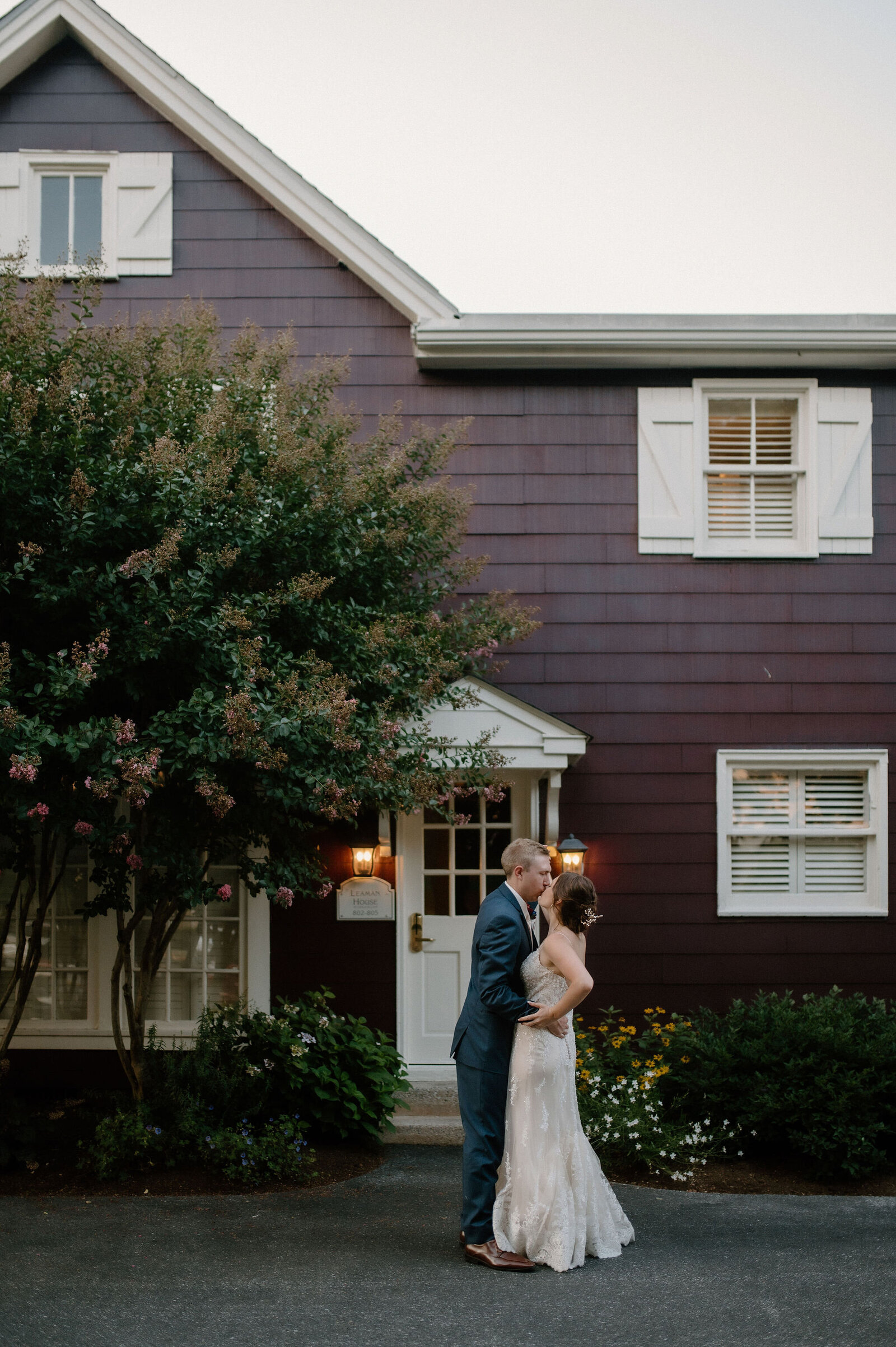 wedding couple kissing in front of maroon house with blooming tree under outdoor porch lights