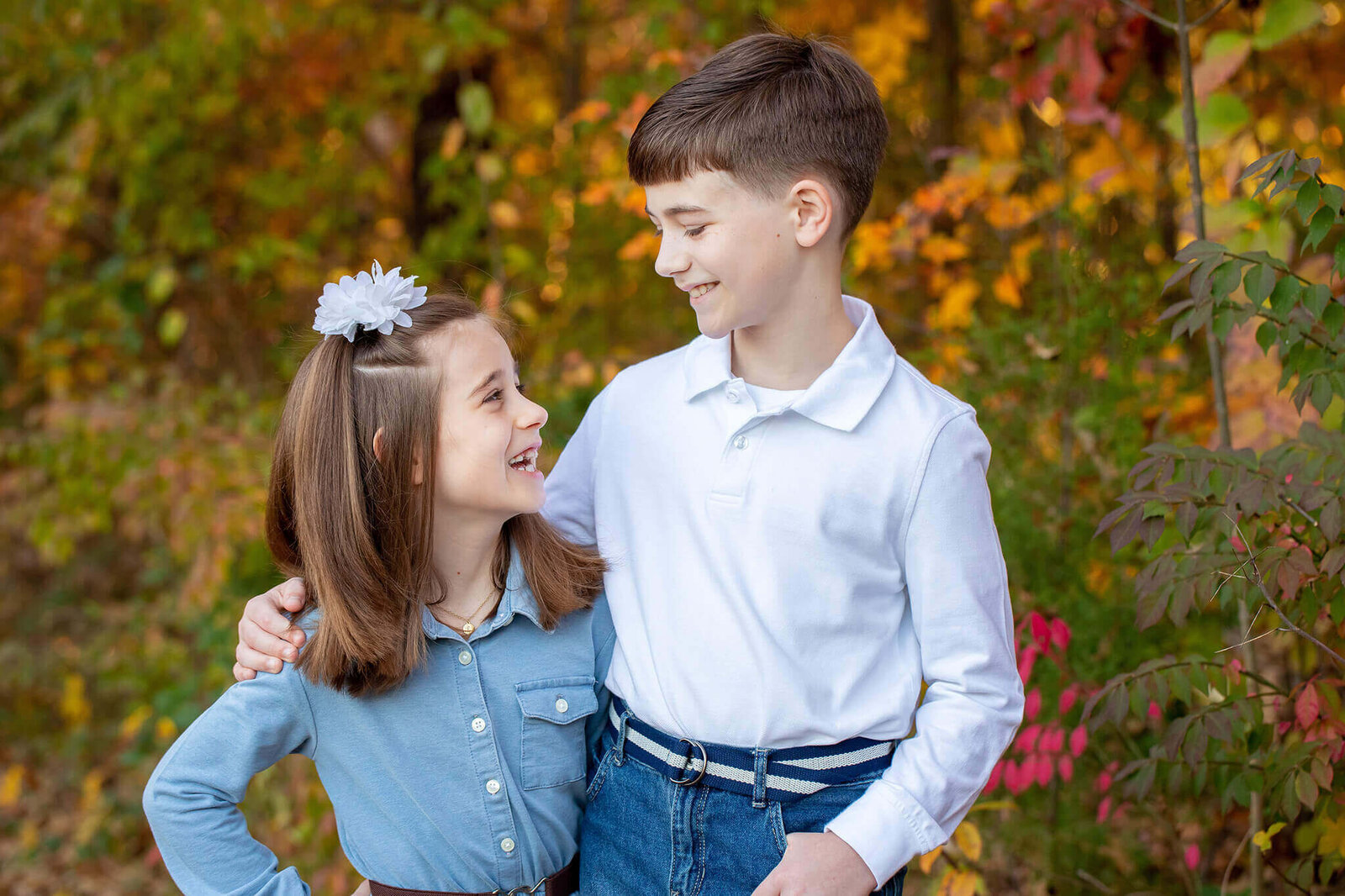 A brother and sister standing among the fall foliage and looking at each other.