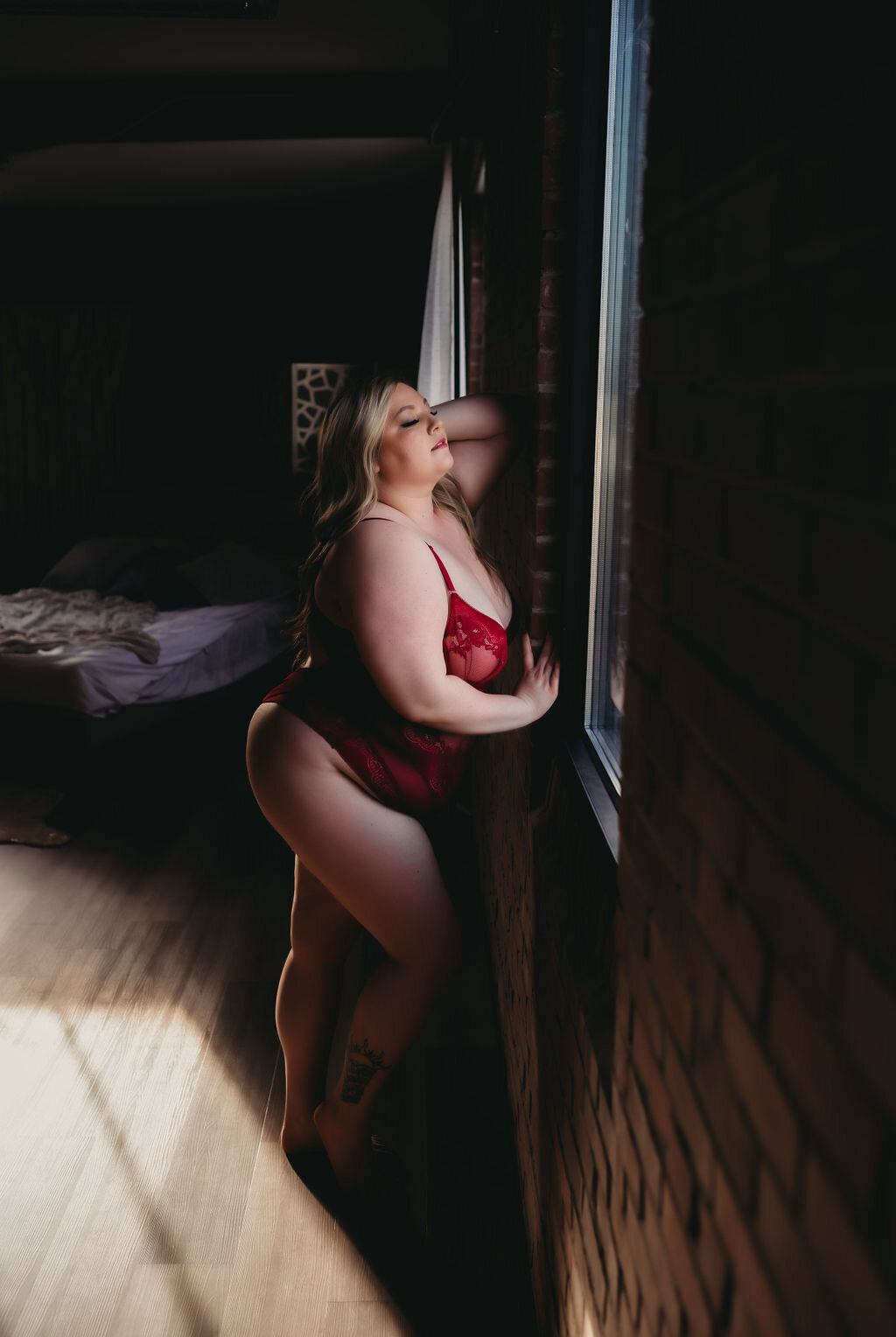 plus size woman in red lingerie leaning against brick wall looking out window with hand in hair