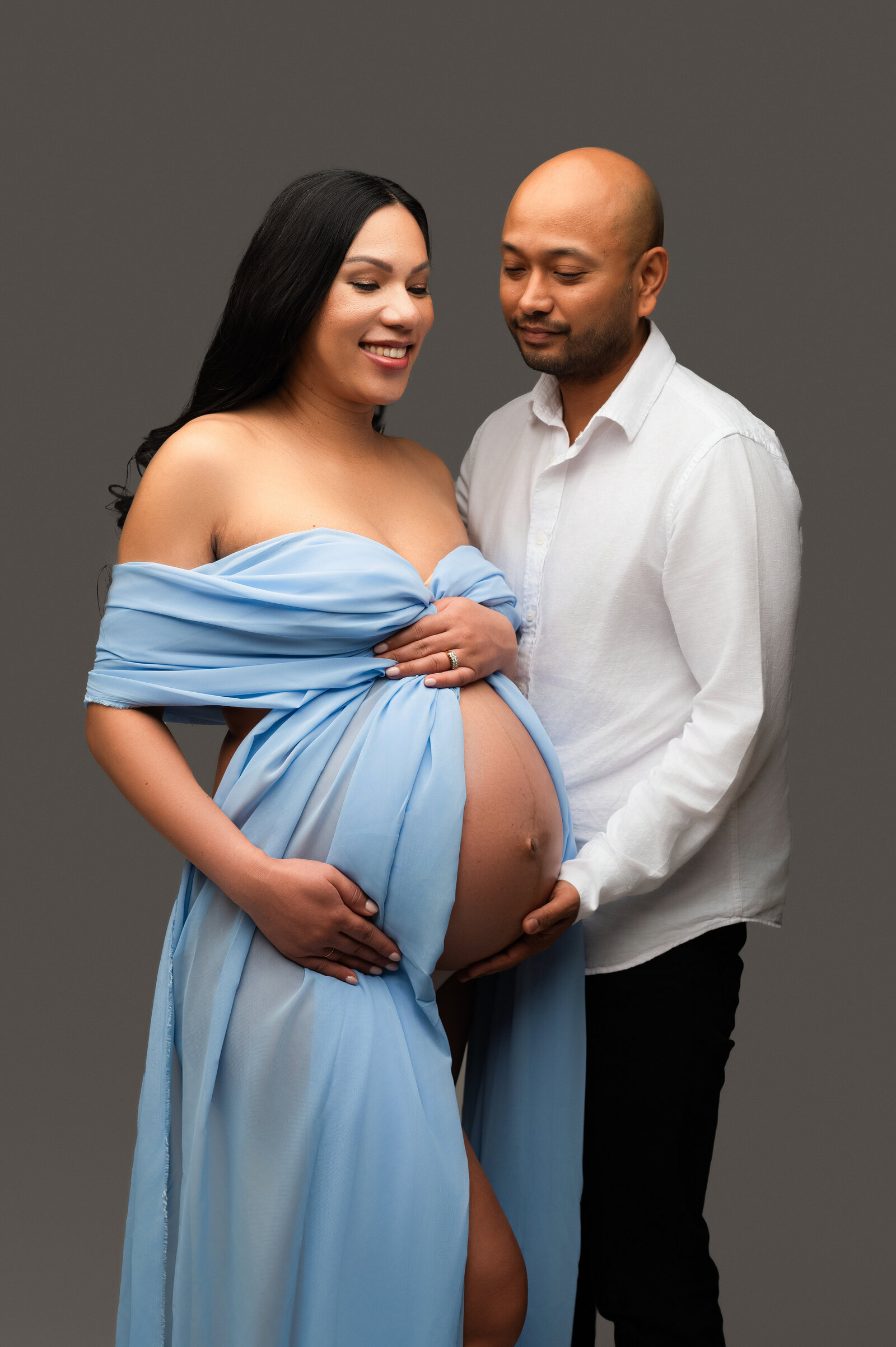 A pregnant woman in a flowing blue dress smiles while holding her belly, captured beautifully at a Charlotte maternity photography studio. A man stands beside her, gently placing his hand on her belly and gazing at her warmly against the plain gray background.