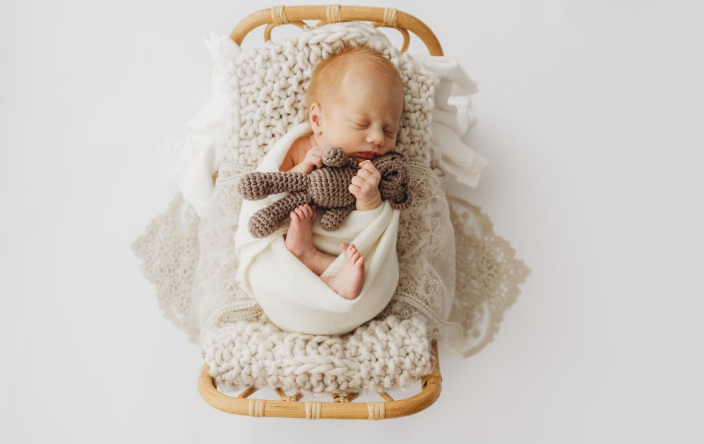 Overhead view of a serene newborn baby in a deep sleep, wrapped in white and holding a tiny knitted teddy, lying in a charming little wooden bed