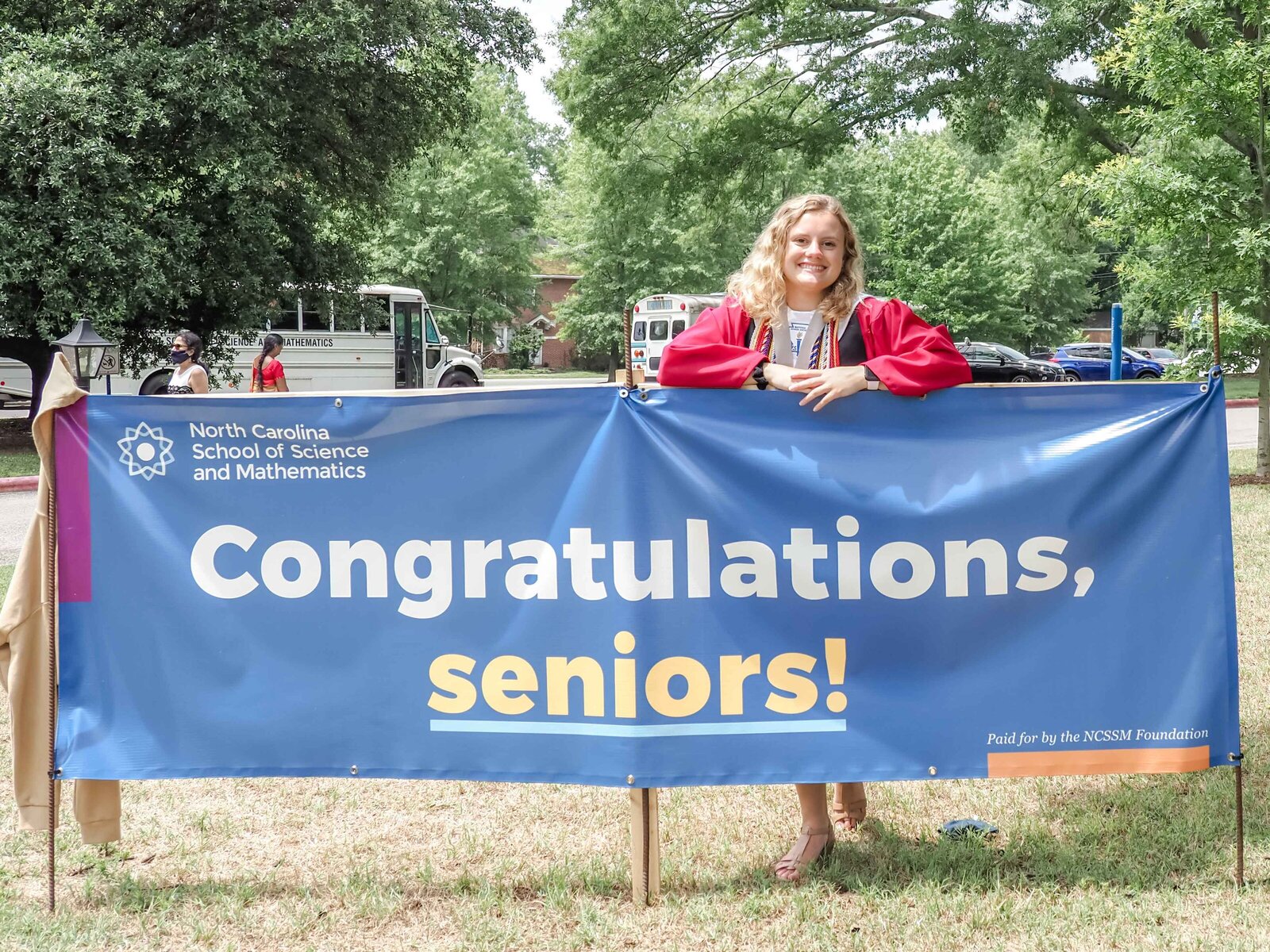 A high school senior in a red graduation gown standing behind a "Congratulations, seniors!" banner at the North Carolina School of Science and Mathematics. This celebratory image captures the joy of graduation, ideal for commemorating academic success.