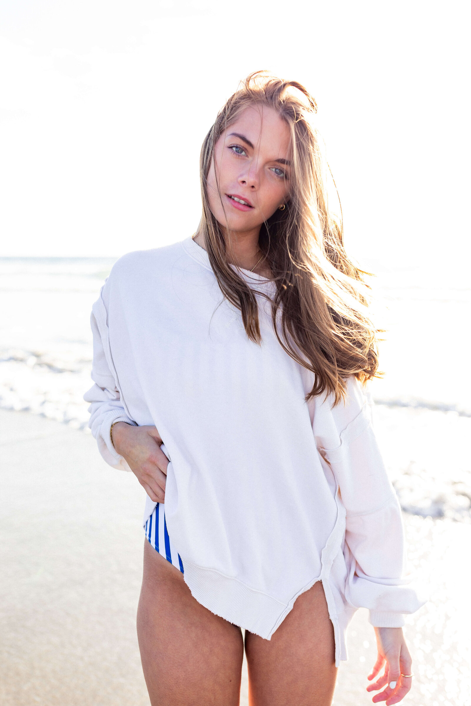 brunette high schooler posing on a beach wearing a white shirt and blue striped swimsuit bottoms