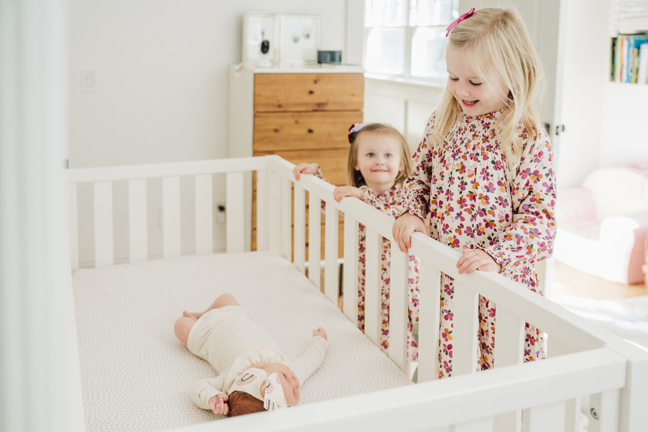 big sisters lean over crib with new baby sister