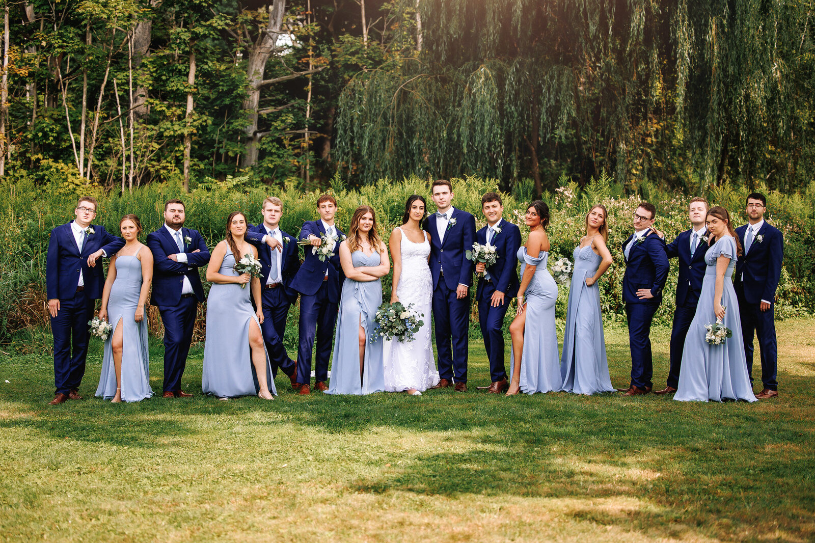 Posing in a casual fashion, a bridal party stands for a photo. The men wearing navy blue suits and the women in long dusty blue dresses. 