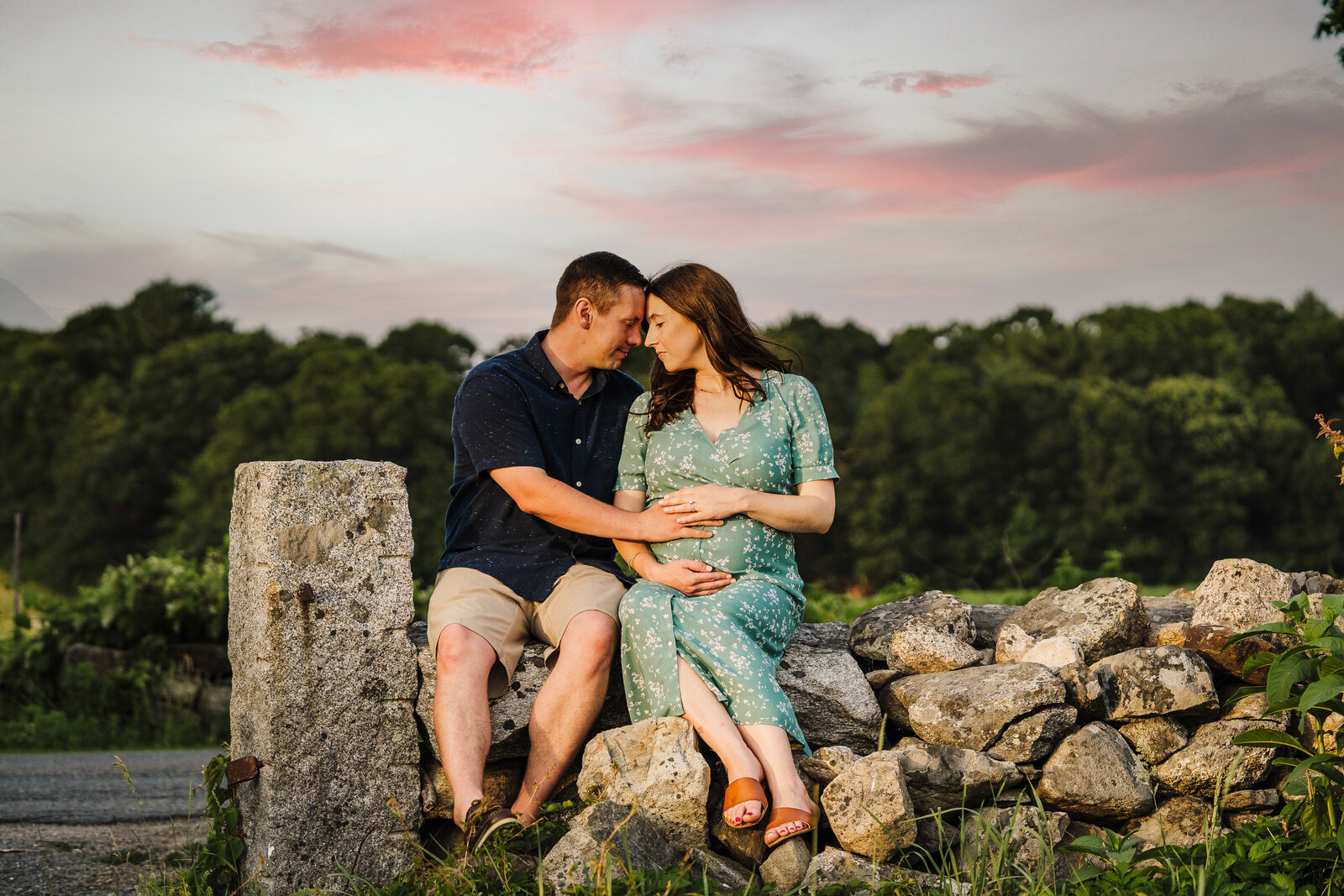 pregnant couple sits on rock wall with pink sunset