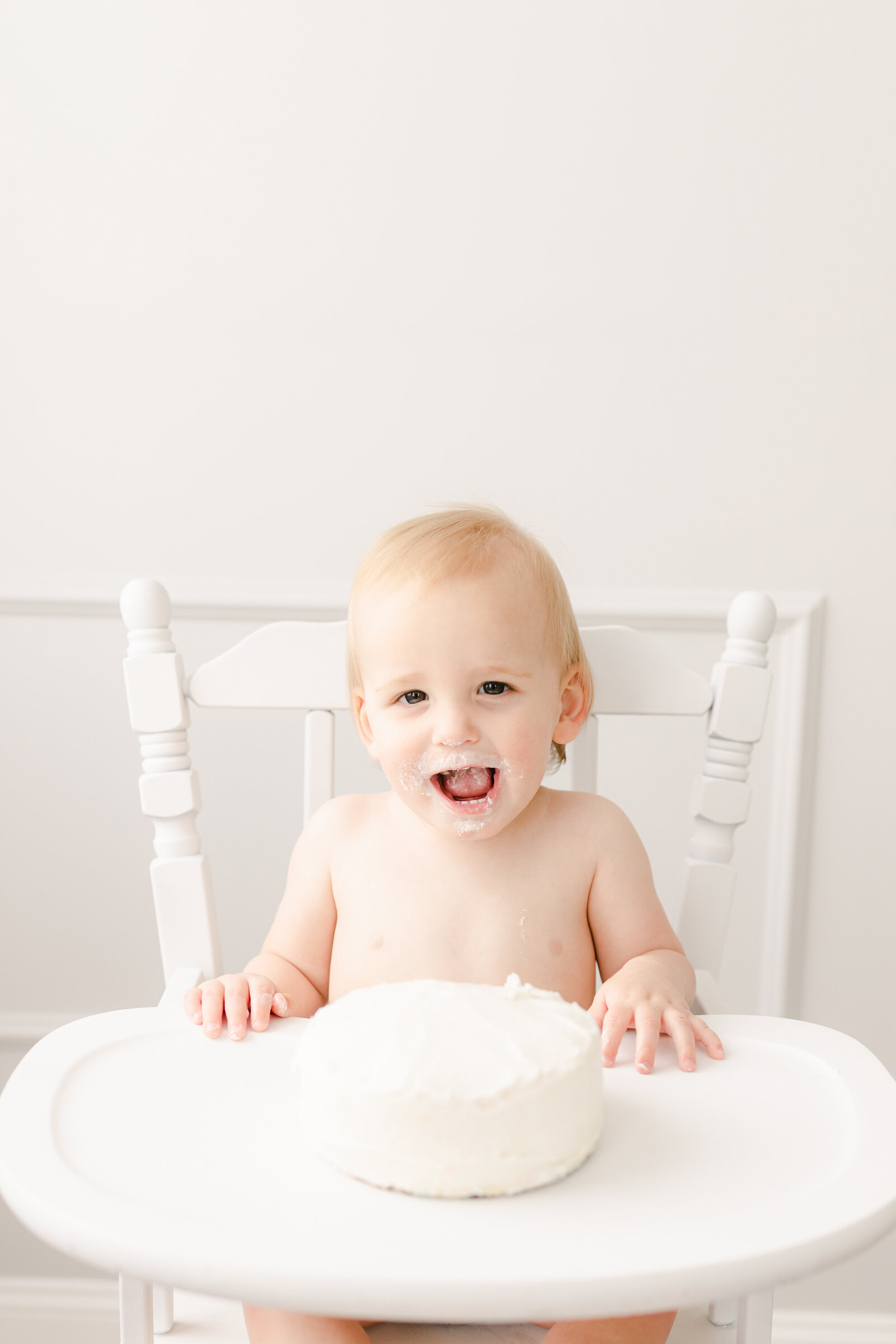 Little boy smiling in his high chair with white frosting on his face and a cake in front of him