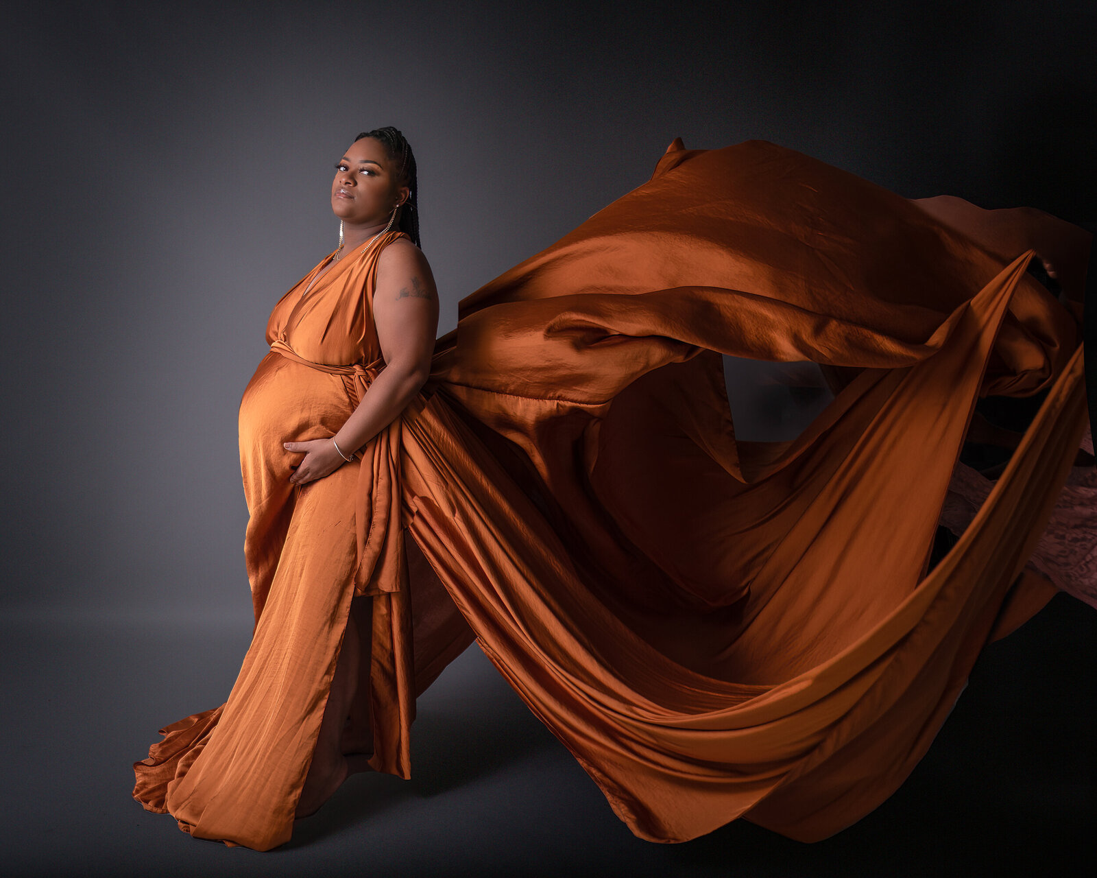 A regal-looking pregnant woman poses in a burnt orange silk gown, her dress billowing around her against a deep, moody backdrop.