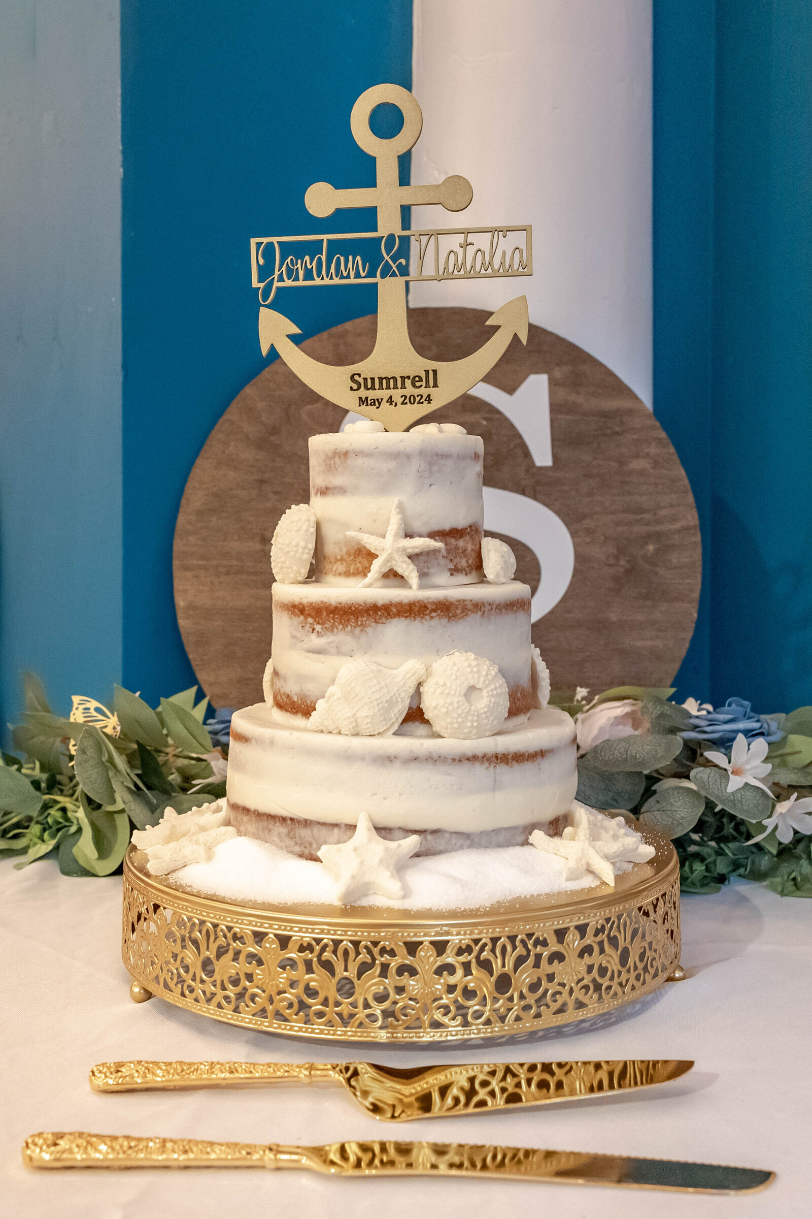 A three-tiered naked wedding cake decorated with starfish and seashells, topped with a custom anchor design and displayed on a gold stand, captured during a nautical-themed Wrightsville Beach, NC wedding.