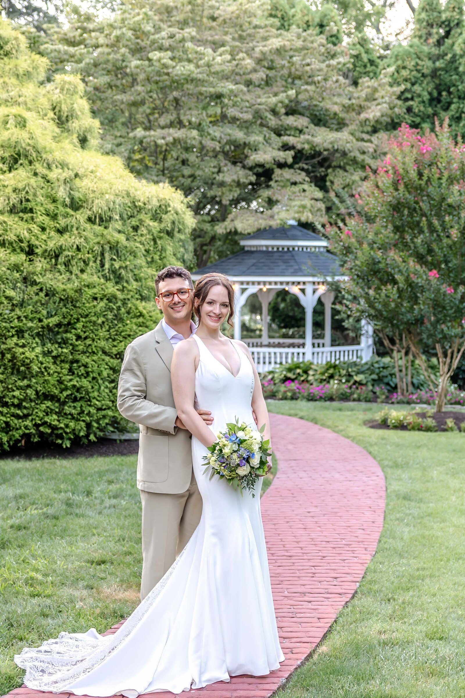 The bride and groom pose on a brick path in a lush garden with a white gazebo in the background, captured during a Wilmington, DE wedding, blending natural beauty with classic romance.