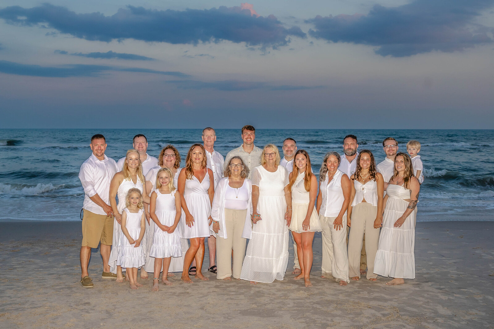 A large family dressed in elegant white outfits poses on the beach at sunset. The ocean and dramatic sky create a stunning backdrop, perfect for North Carolina family photography, capturing timeless and elegant family gatherings.