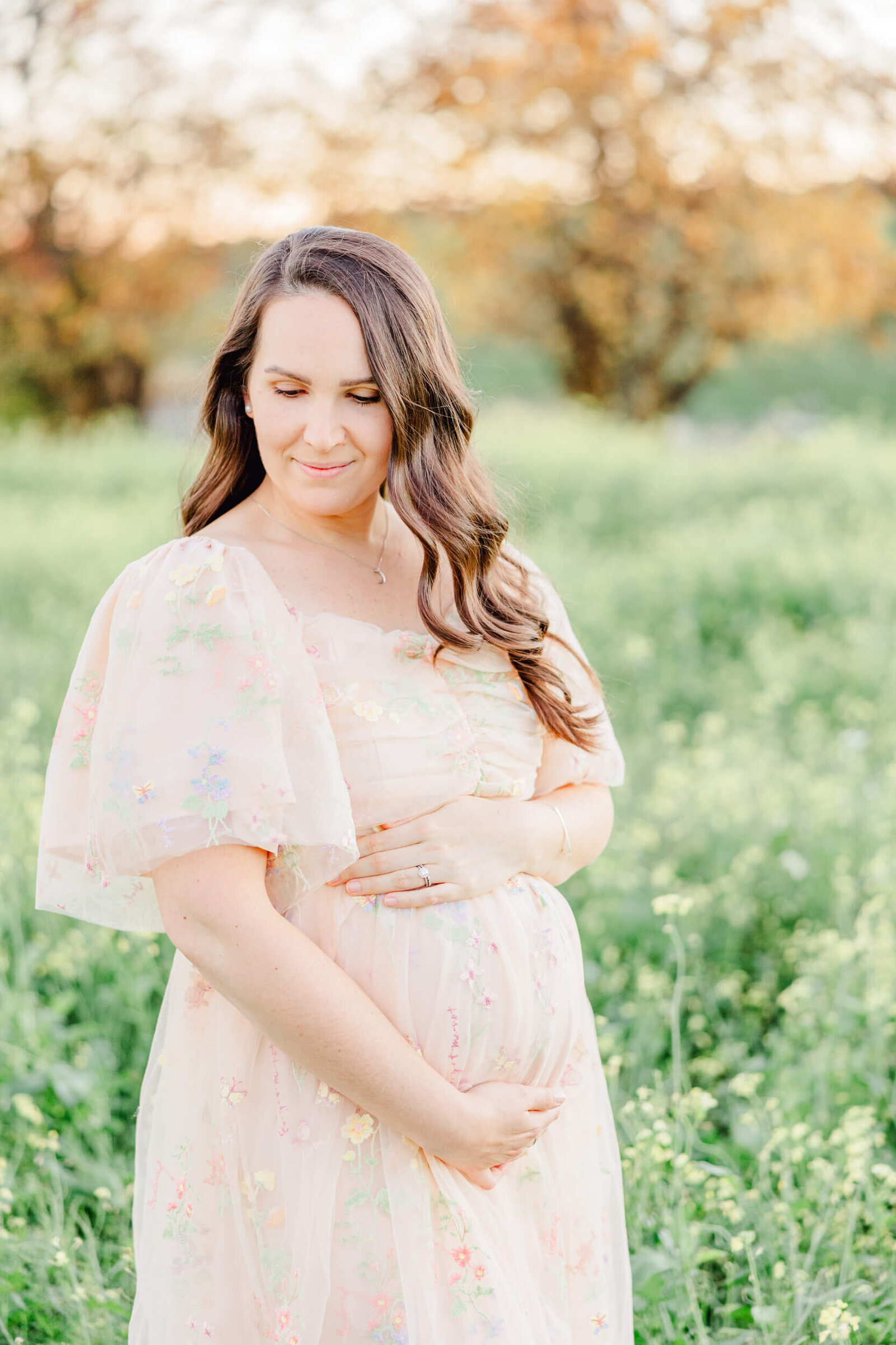 Pregnant mom in a soft pink dress with delicate floral embroidery cradles her bump and smiles softly to the side