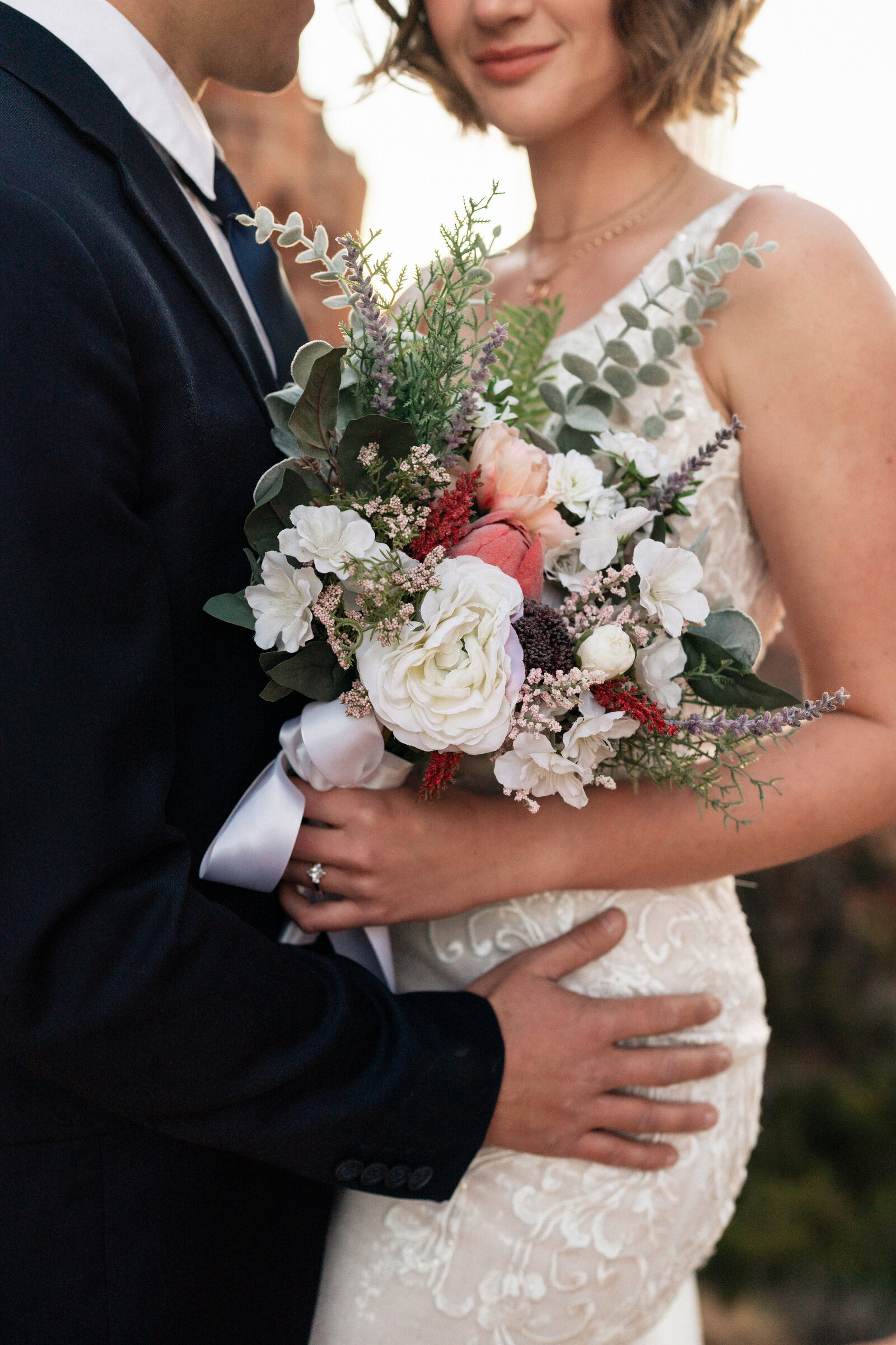 Close-up of bride holding a vibrant floral bouquet with white, pink, and lavender blooms during her elopement at Cathedral Rock in Sedona, Arizona, with the groom’s hand gently placed on her waist.