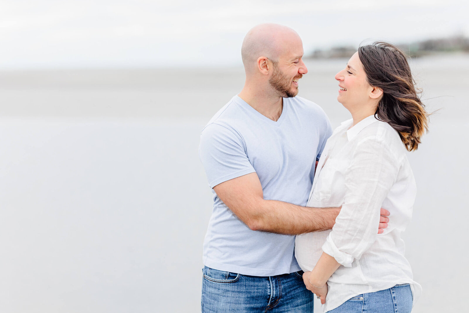 Man hugs his pregnant wife as they smile in front of the ocean