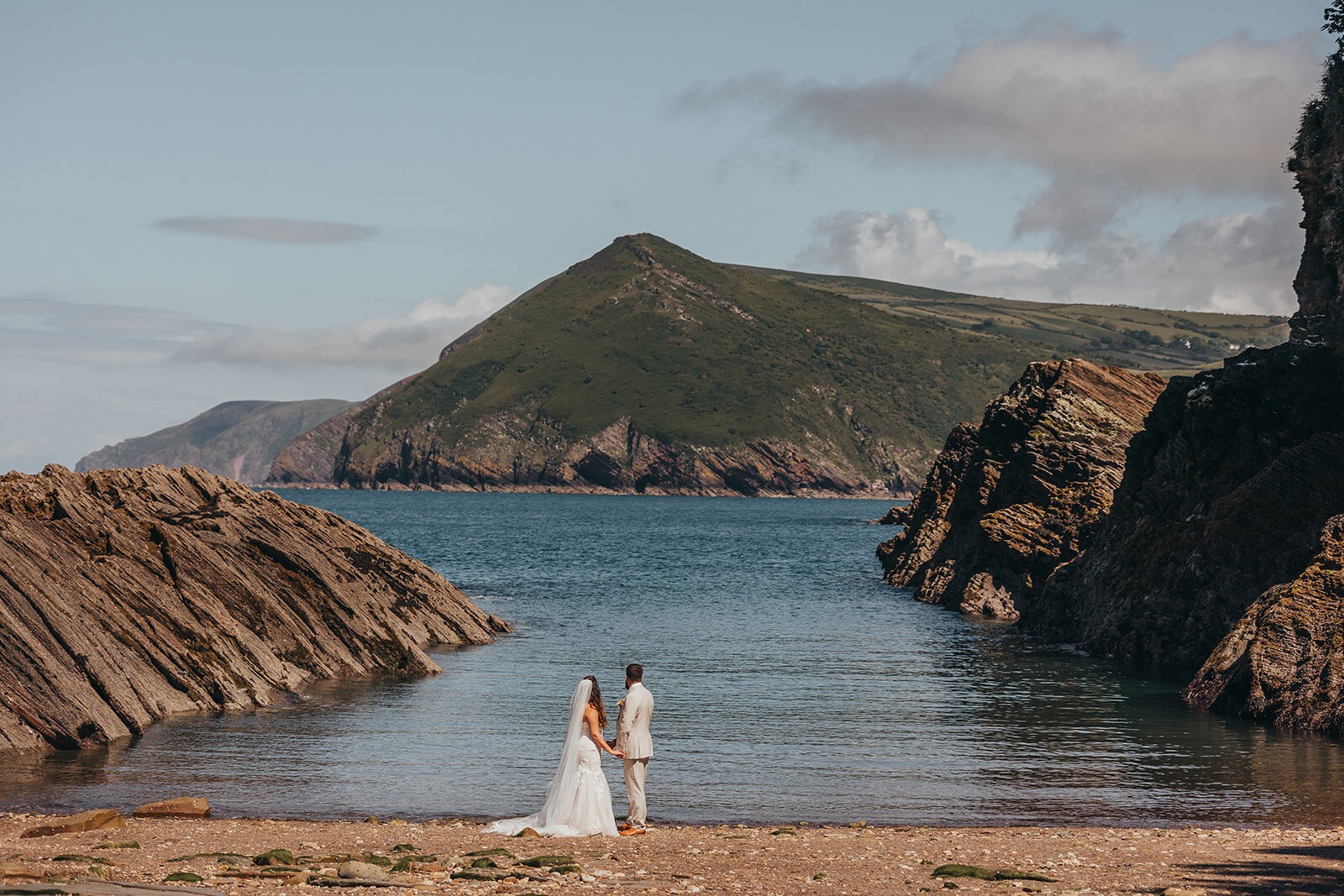 bride and groom stand on a beach looking out to sea.