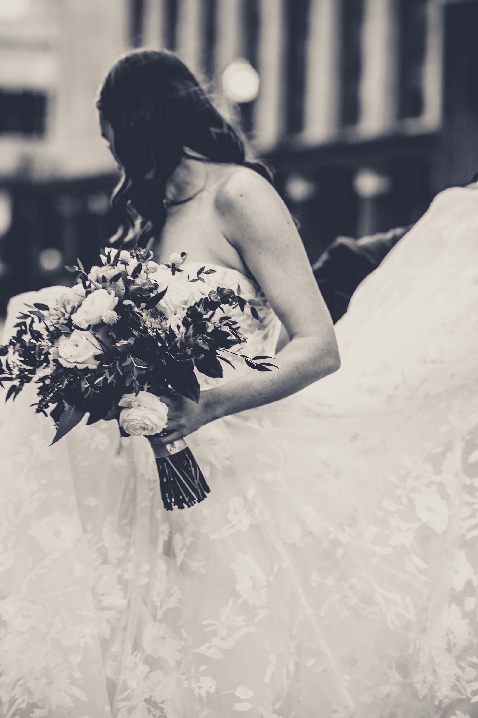 Capture the timeless elegance of this black and white photograph featuring a radiant bride holding her beautiful bouquet. The image highlights a moment of bridal grace, as her exquisite gown is delicately fluffed, adding a dynamic and flowing element to the scene. Perfect for those who appreciate the classic charm of monochrome photography, this photo beautifully combines the purity of the bride's joy with the intricate details of her wedding dress and floral arrangement.