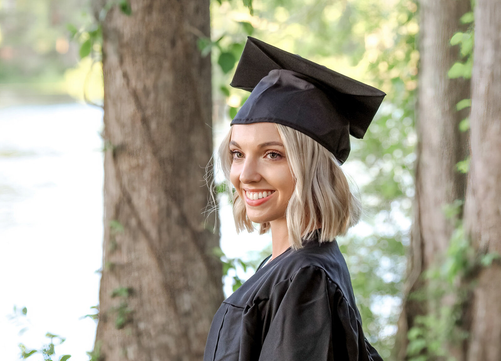 A high school graduate in a cap and gown smiling brightly with a wooded background in Wilmington, North Carolina. This portrait captures the joy of graduation, ideal for those seeking natural and professional senior photography.