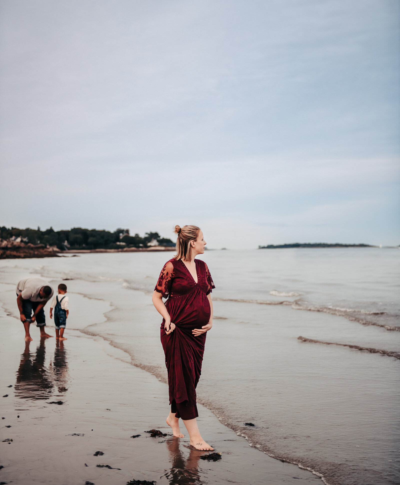 Maternity Photographer, Pregnant woman wearing a dress walking on a beach while a man and little boy play behind her.