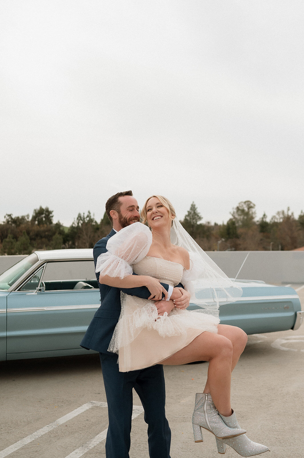 groom holding up bride with blue vintage elopement car in background