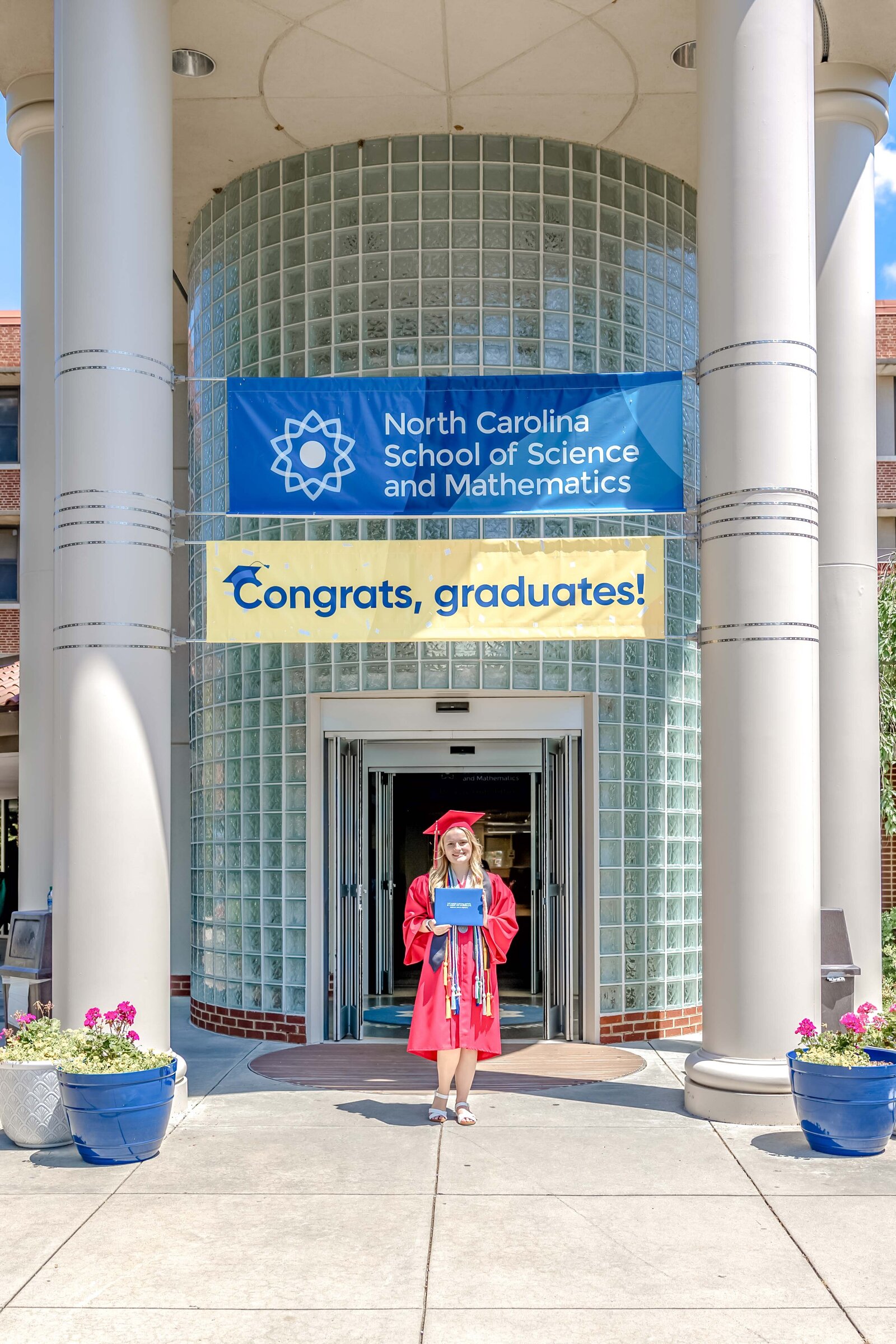 A senior in a red gown holding a diploma in front of the North Carolina School of Science and Mathematics with a congratulatory banner. This celebratory photo captures graduation success, ideal for capturing meaningful senior photography.
