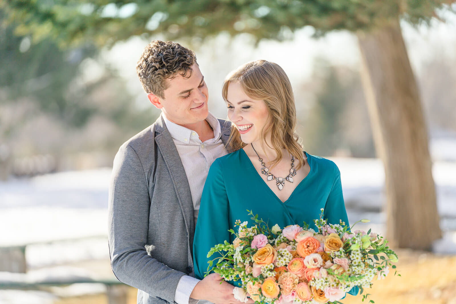 A man in a white button-up shirt and a gray blazer holding his his fiancee wearing a long sleeved dark teal dress, holding a bouquet of orange and pink roses while standing in a snow covered park in Salt Lake City
