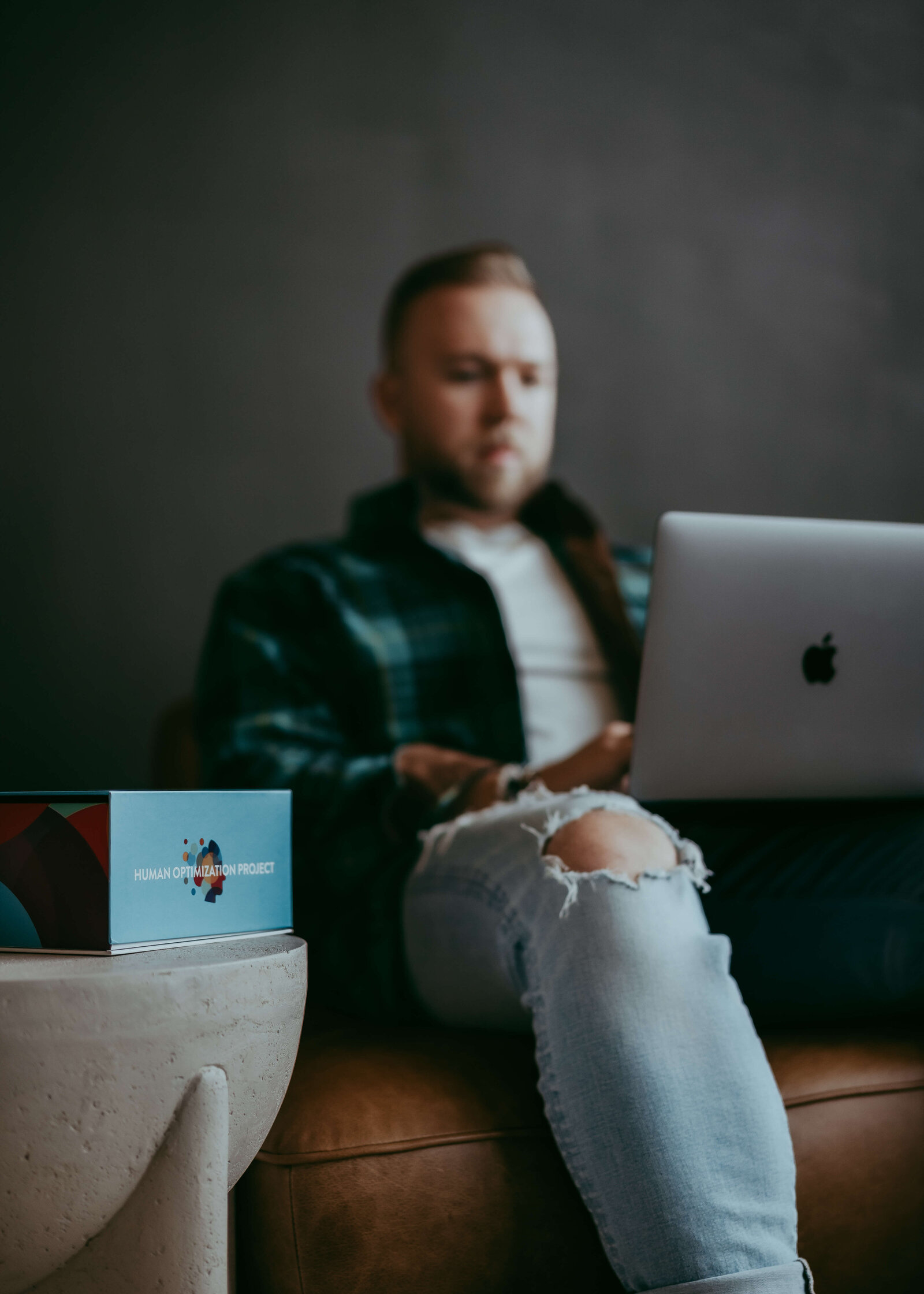 A man sits wearing a green and navy unbuttoned shirt while working on his MacBook during a branding session for his supplement company that product is sitting on a table near him.