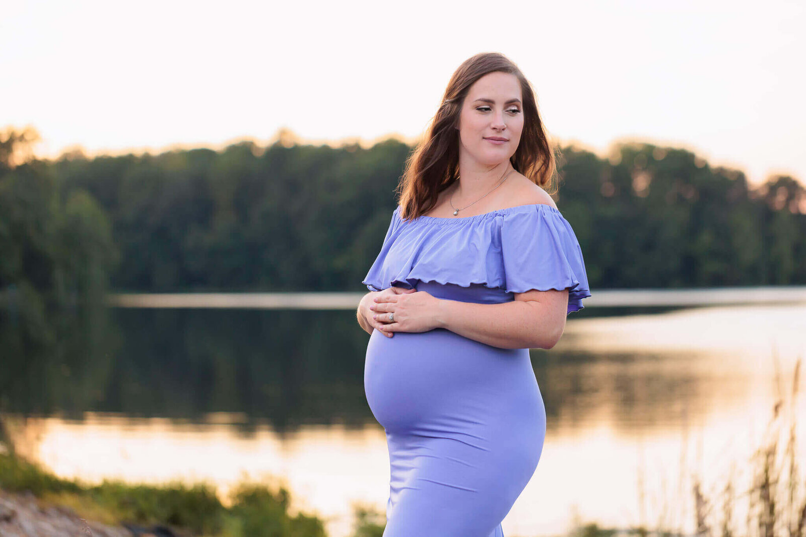 A pregnant woman in a purple dress posing in front of a lake in Fairfax County.