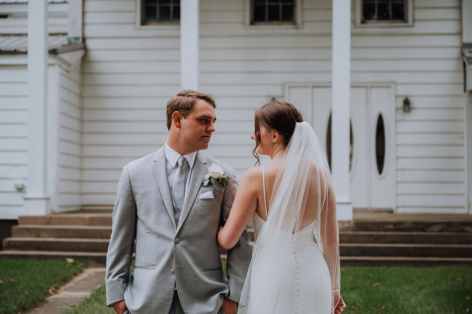 Groom facing camera and bride facing away with arms interlocked and the couple looking at one another