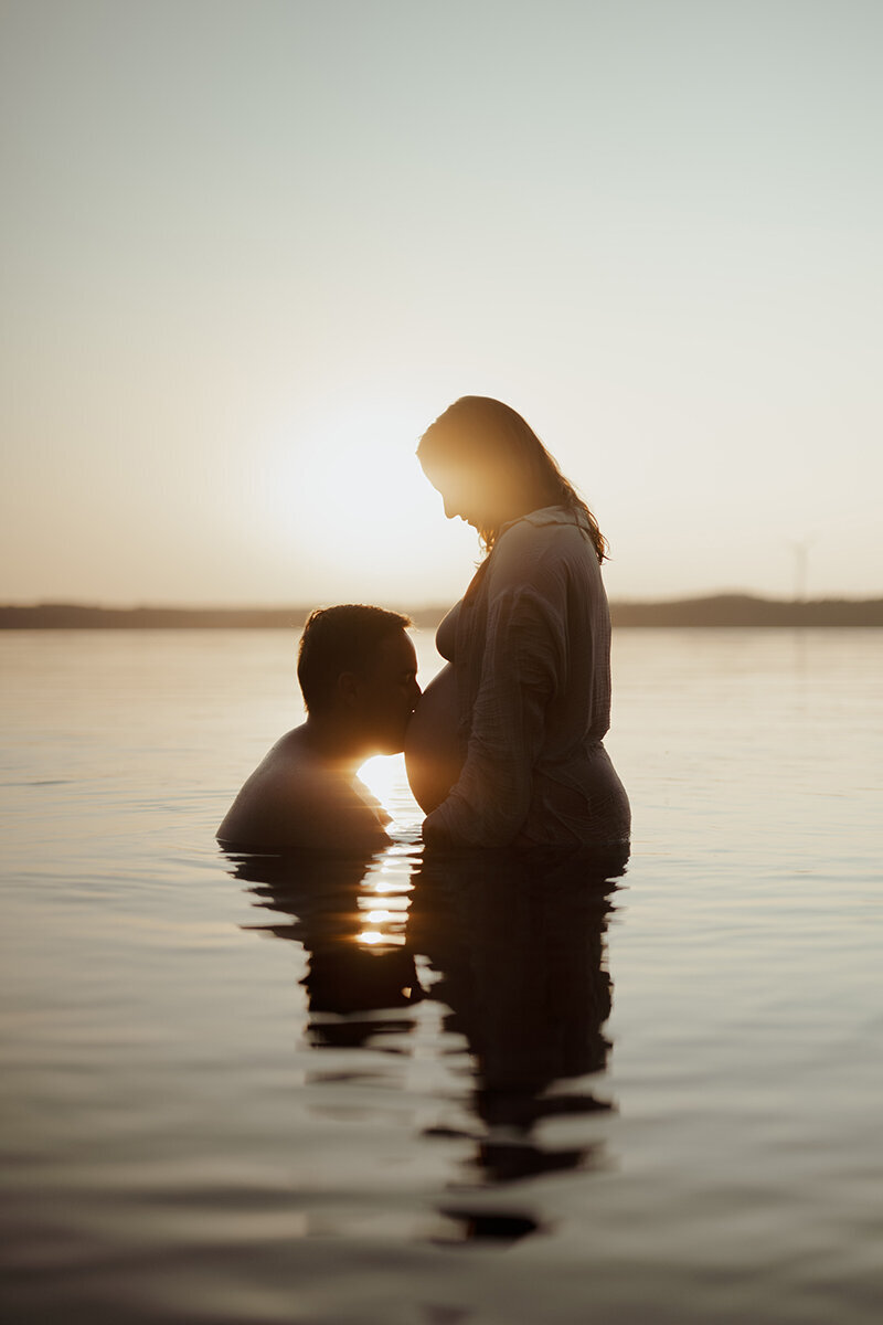 Gravid kvinna står i vatten med sin man som kysser henne på magen i solnedgång