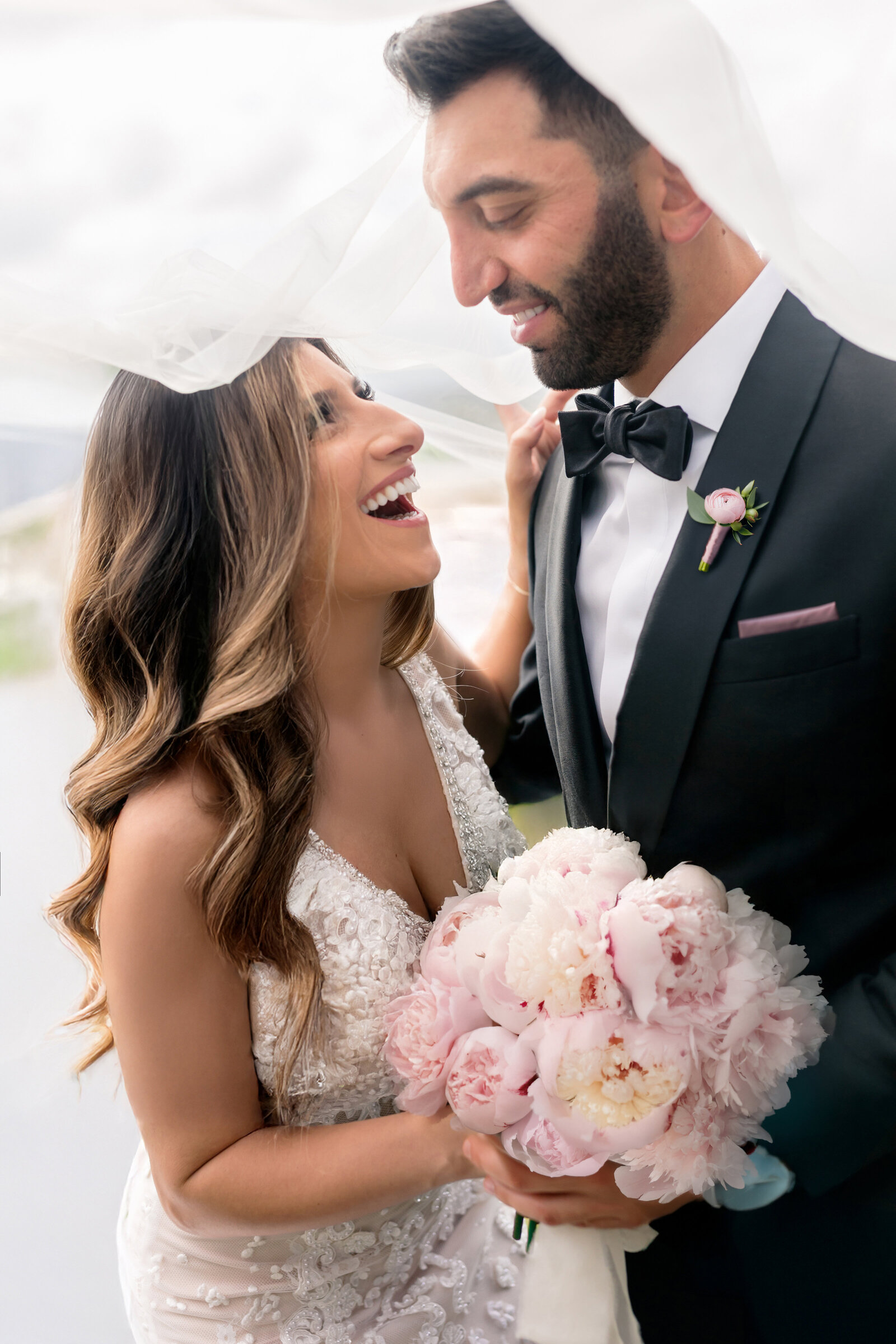 A bride in a white dress and a groom in a black tuxedo share a joyful moment under a veil. She holds a bouquet of pink and white flowers, smiling up at him. They stand against a soft, blurred outdoor background.