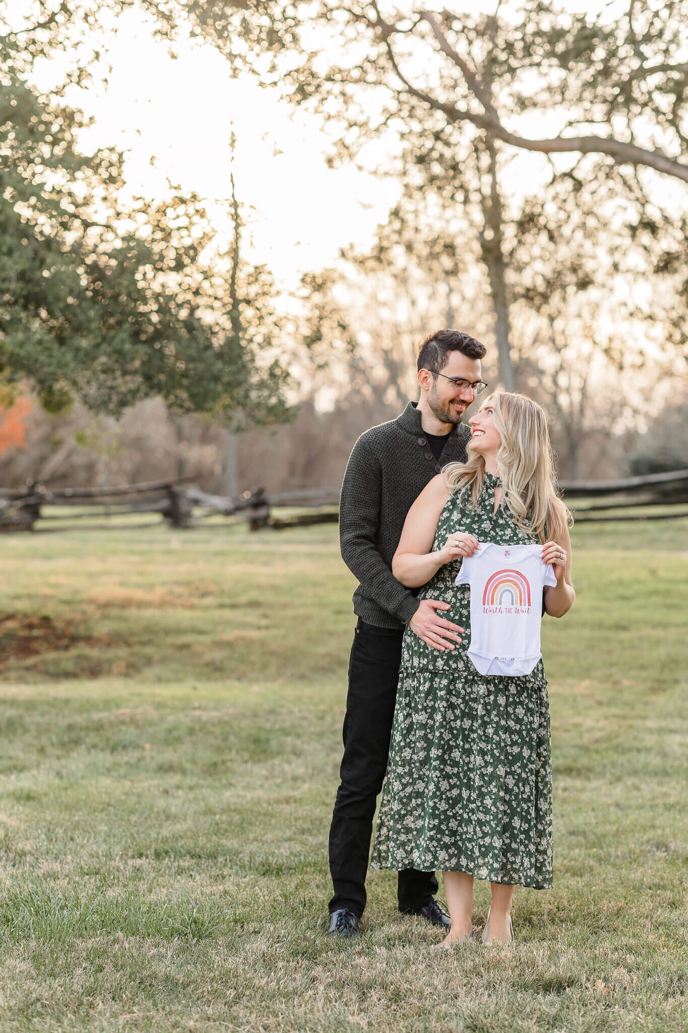 A pregnant woman holding a onesie in front of her while her husband hugs her from behind.