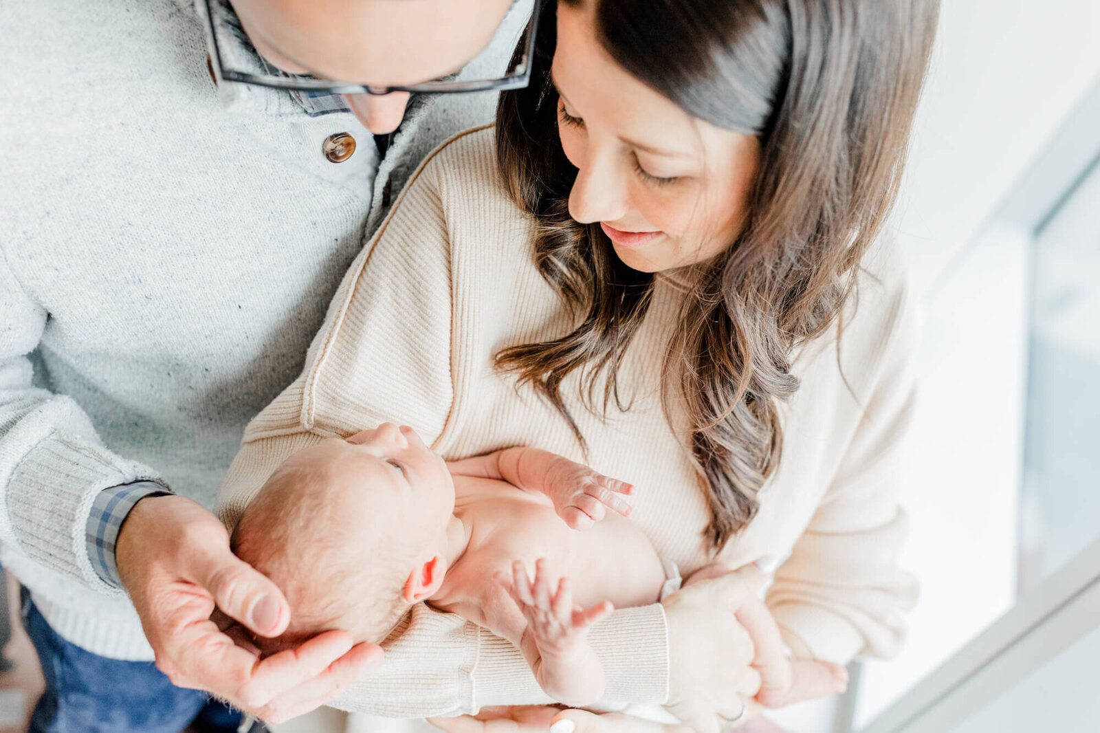 Taken from above, mom and dad stand together and gaze at their newborn, with mom holding him and dad cupping the baby's head with his hand