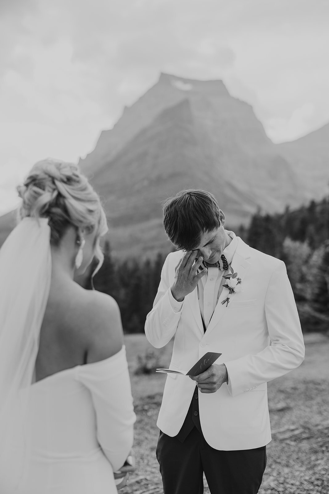 A groom wipes away tears while reading vows to his bride against a mountain backdrop in Glacier National Park, captured in black and white by Sydney Breann Photography during their emotional elopement ceremony.