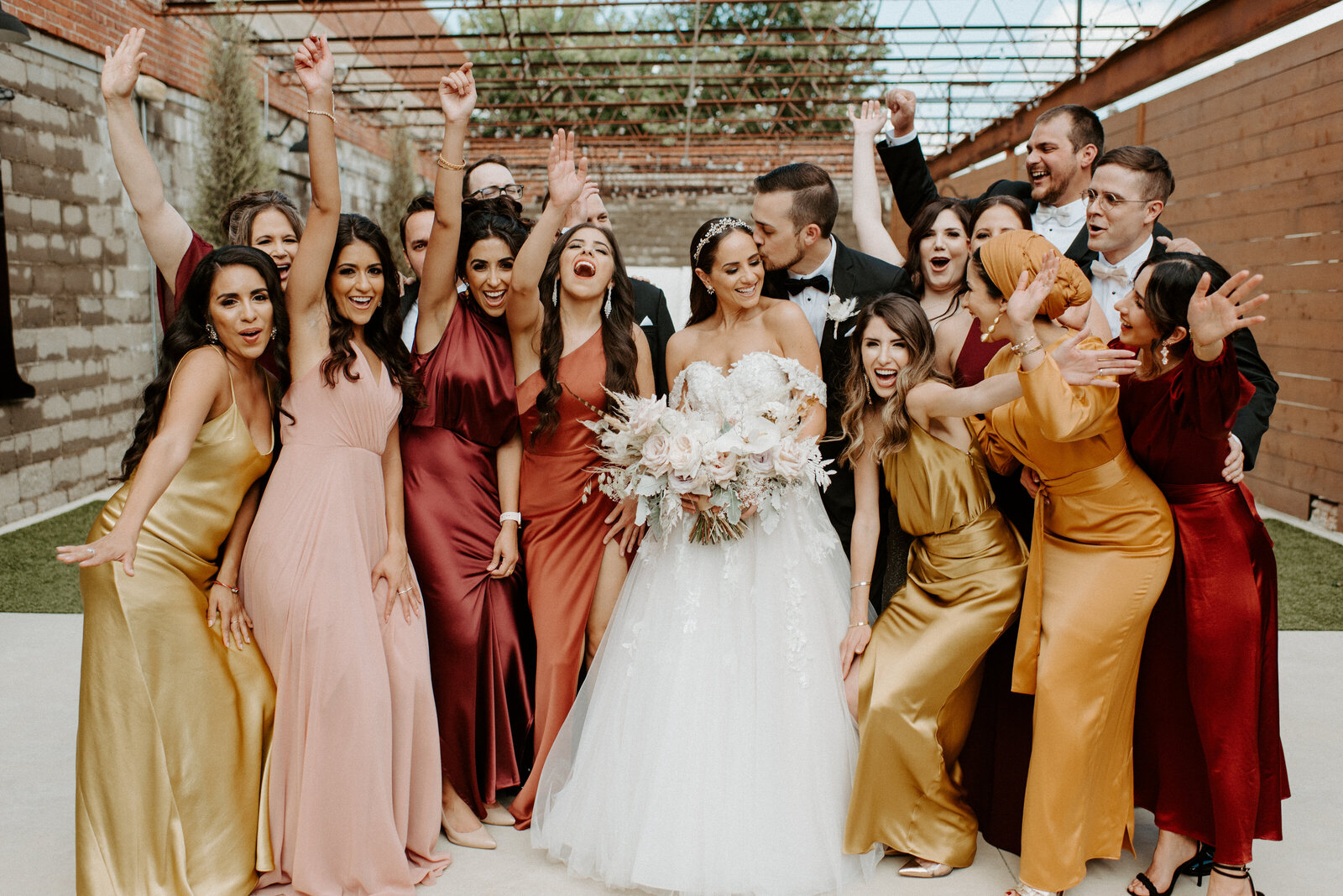 A group of bridesmaids and groomsmen celebrates around a bride and groom as he kisses her cheek at The Hudson event venue in Wichita, Kansas. The bride holds a beautiful bouquet,  the women are wearing different colors of dresses, and the men are waring tuxedos.